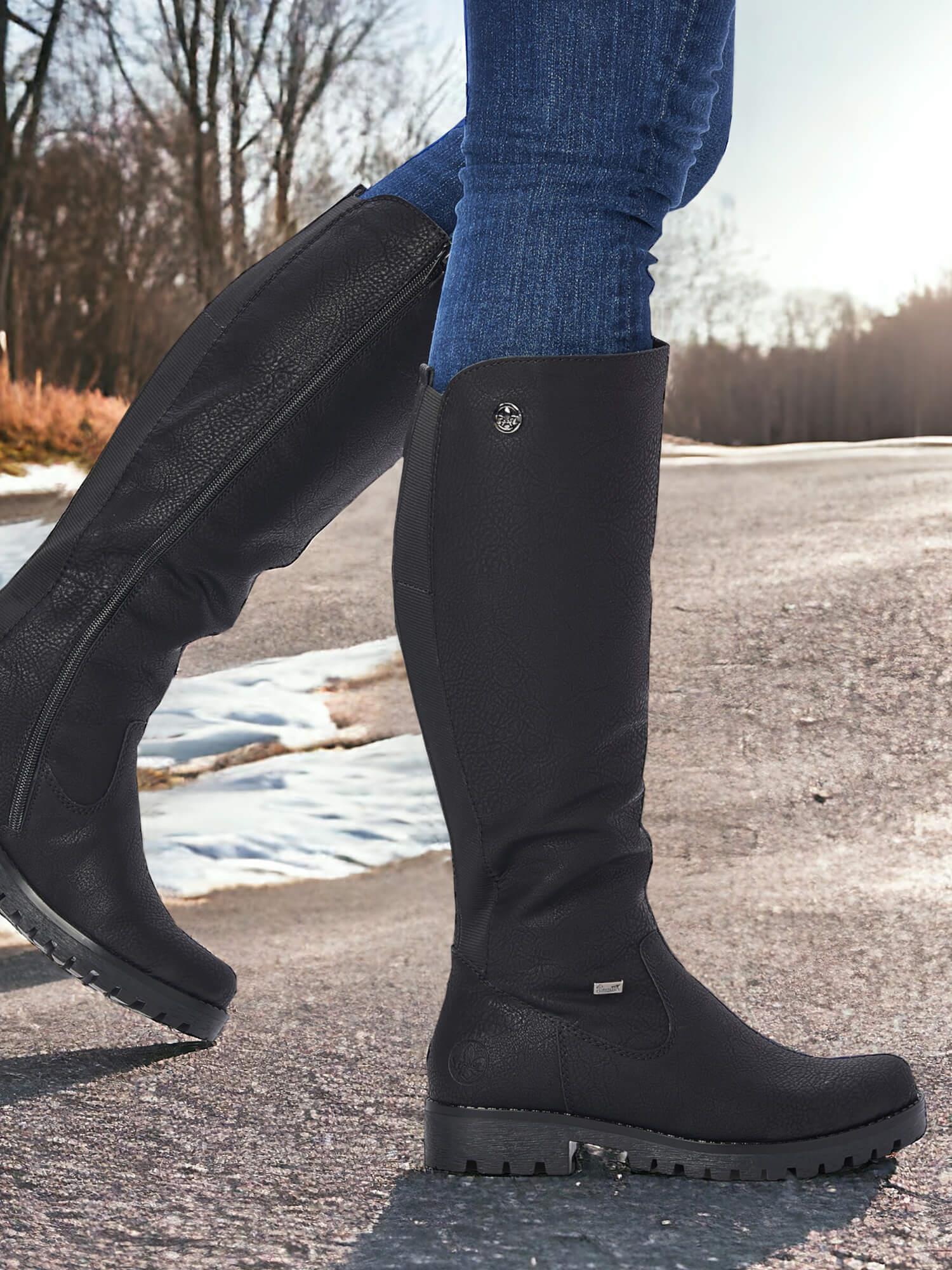 En person iført 07856 Rieker Comfort-støvler ullfòr fra Rieker Comfort & Style og blå jeans går langs en snødekt, asfaltert vei med nakne trær under en solfylt himmel.