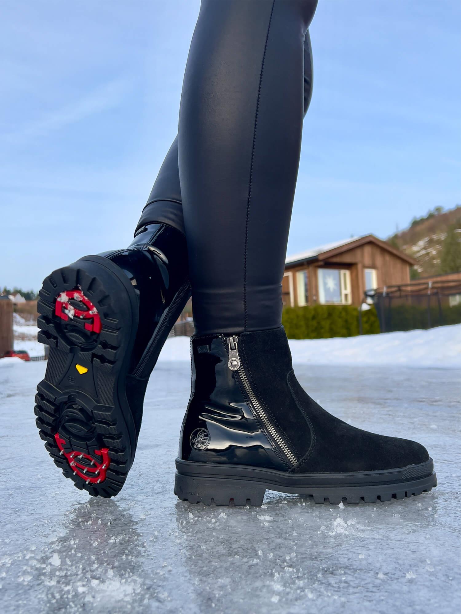 Nærbilde av en person iført Rieker Comfort & Style 05389 Varmfòret med brodder Dame over blanke, svarte leggings, stående på isete underlag utendørs, med hus og trær i bakgrunnen under en klar himmel.