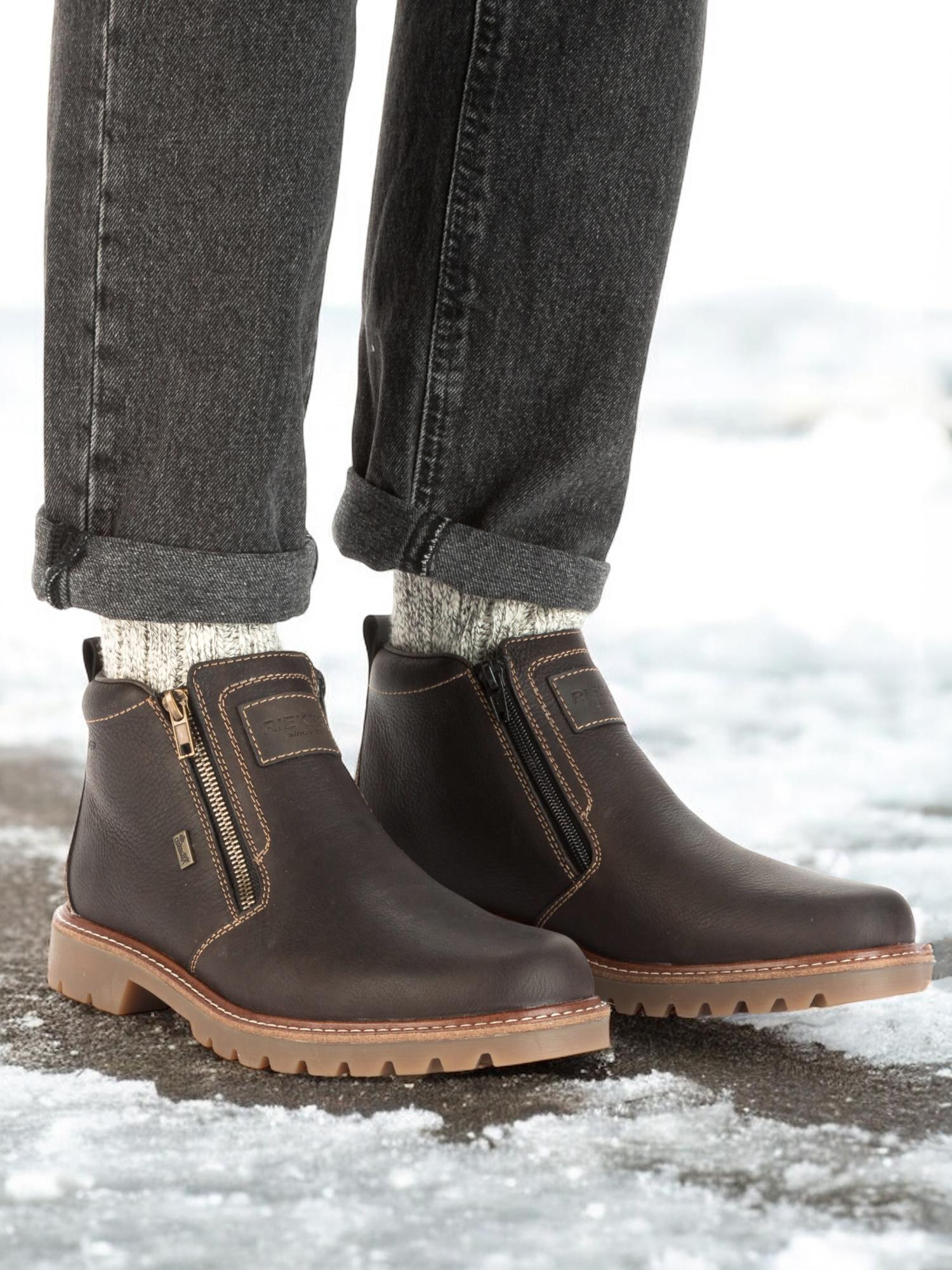 En person iført 04984 Rieker Leather Fiber-Grip ullfôret ankelboots fra Rieker Comfort & Style, med sidelås, grå oppbrettede jeans og lysegrå sokker, står på snø - ideelt for deg som vil ha vannavvisende vintersko.