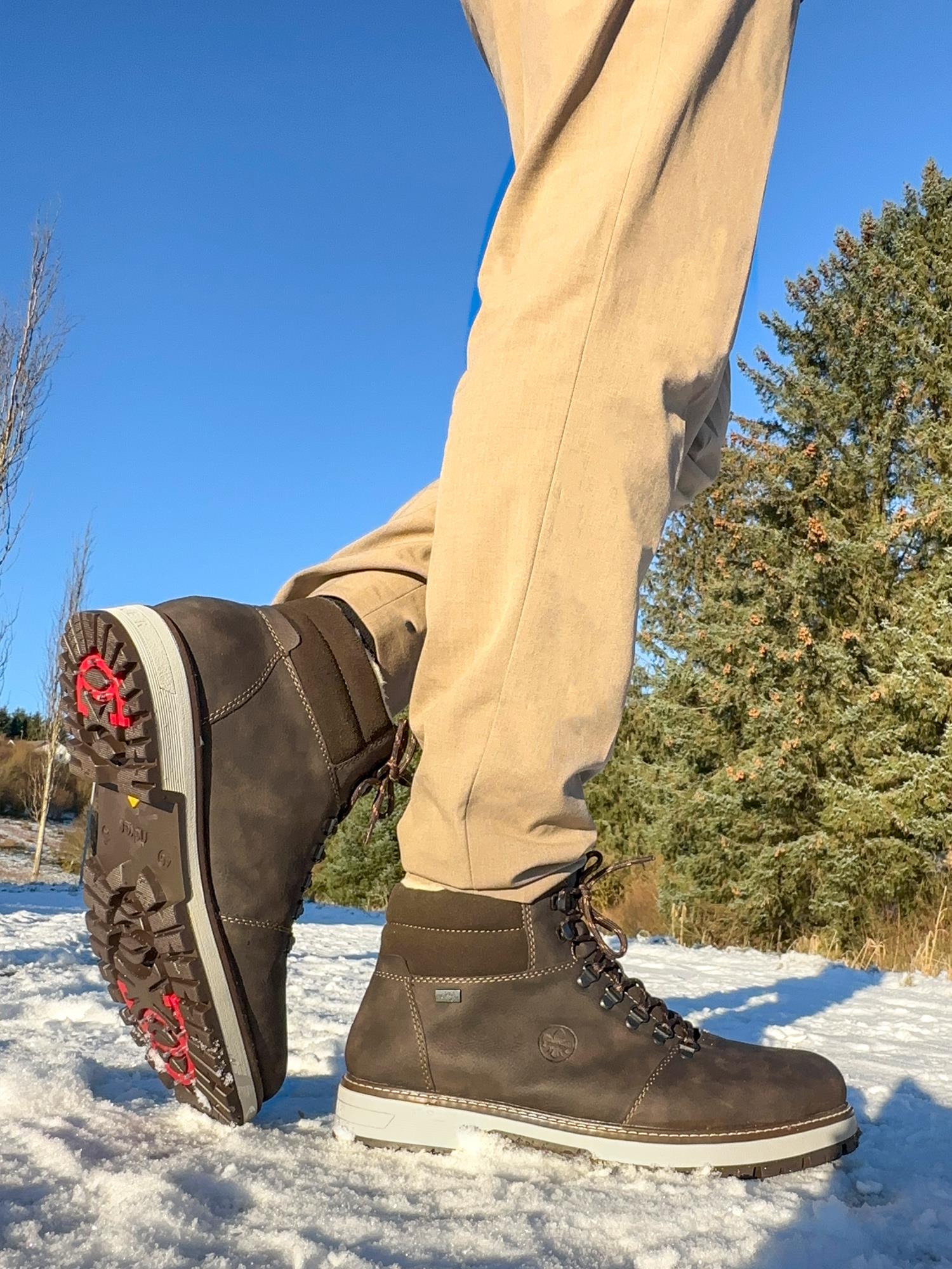 En person iført 02588 Rieker Brown ullfôr TEX-membran med brodder fra Rieker Comfort & Style og beige bukser står i snø og løfter den ene foten. Grønne trær og en klarblå himmel er synlig i bakgrunnen.