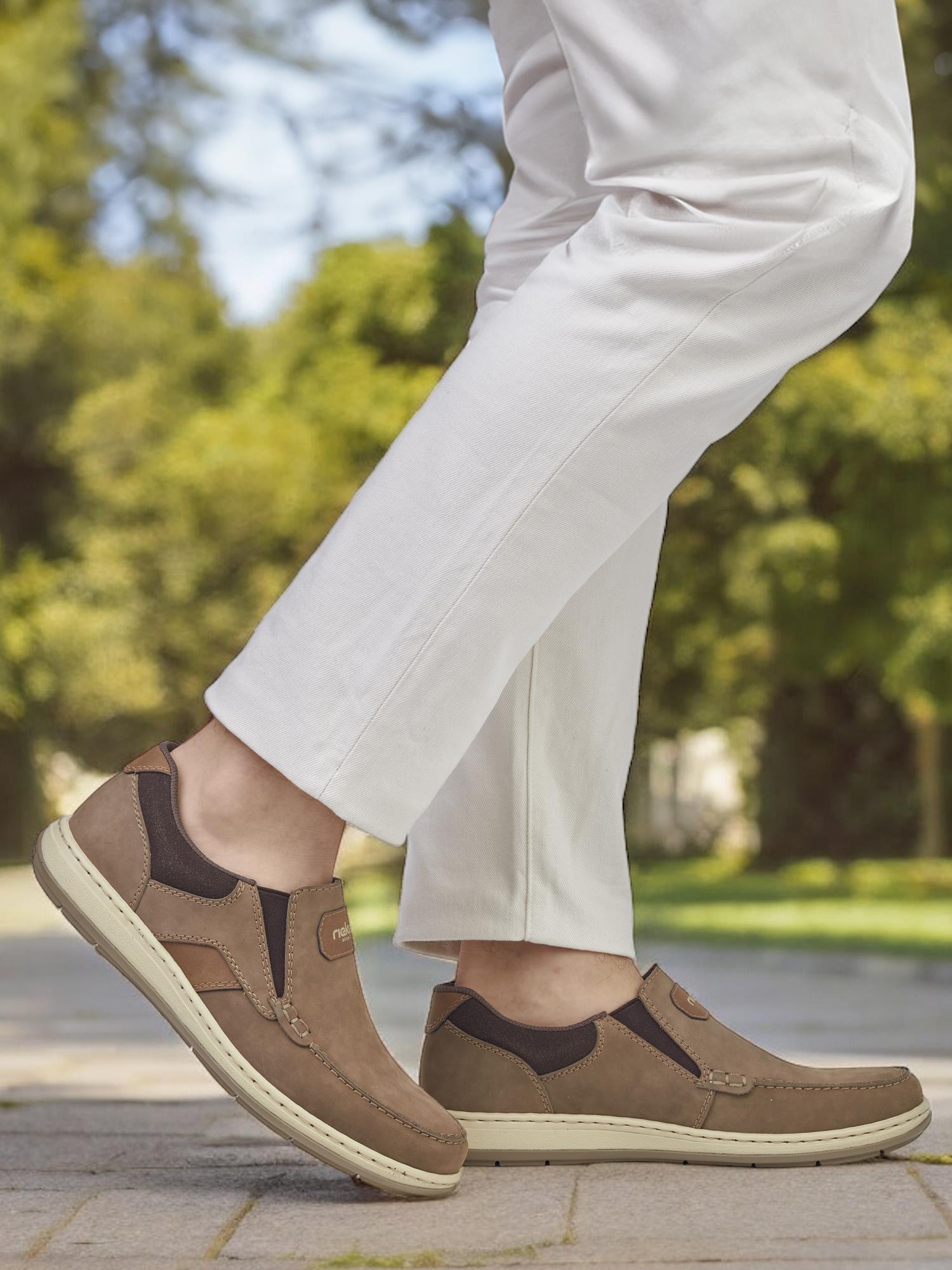 Iført off-white bukser og 07530 Rieker Slip in Loafer Wide Fit Brown fra Rieker Comfort & Style, nyter denne moteriktige mannen hverdagskomfort mens han spaserer utendørs langs en sti omkranset av frodige grønne trær og gress.