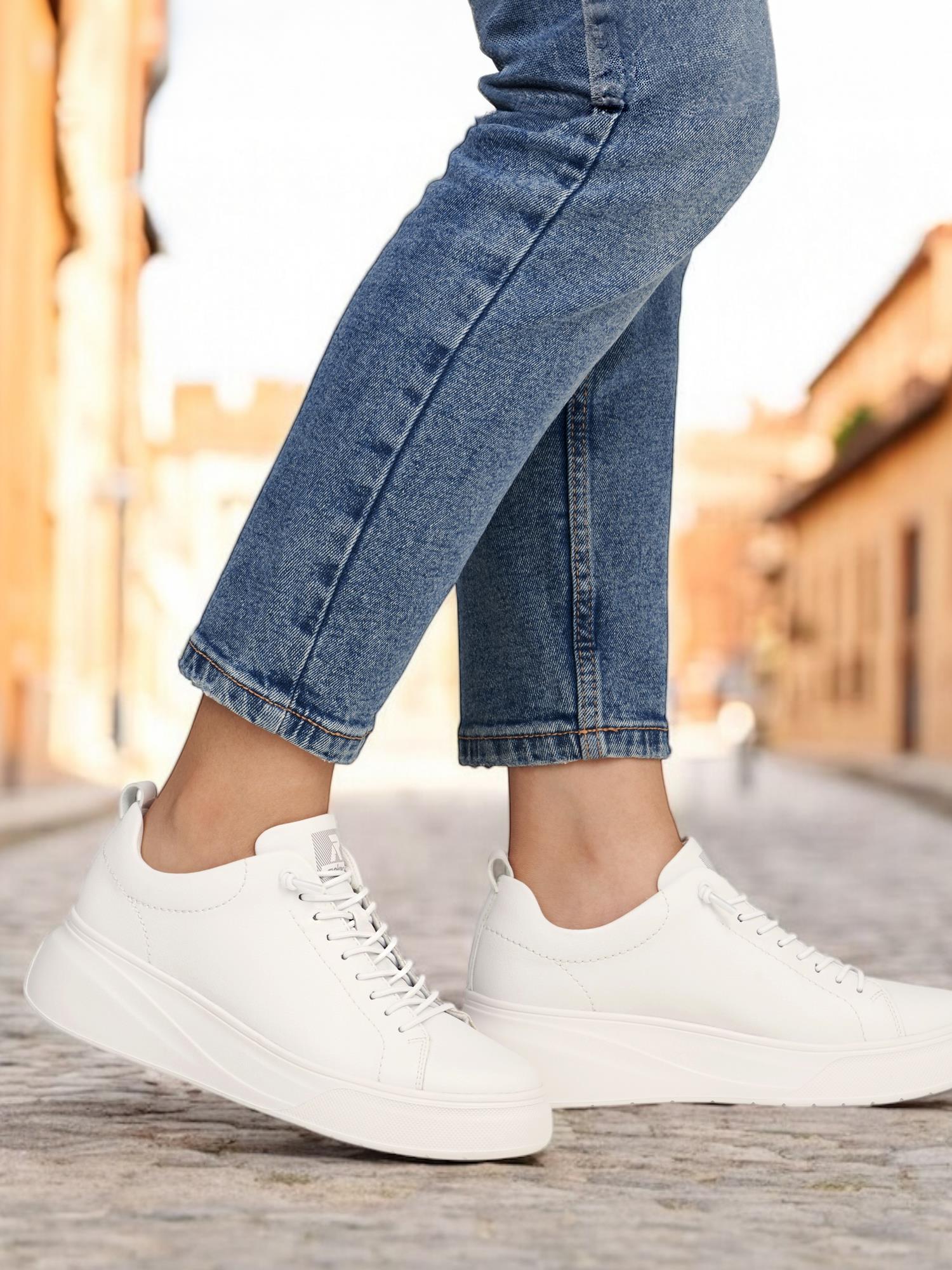 En person i lyseblå jeans og Rieker Sports 03538 Summer Ease White Leather joggesko står på en brosteinsgate med solbelyste, uskarpe bygninger - og viser frem stilig og komfortabelt fottøy som er perfekt for sommerdager.