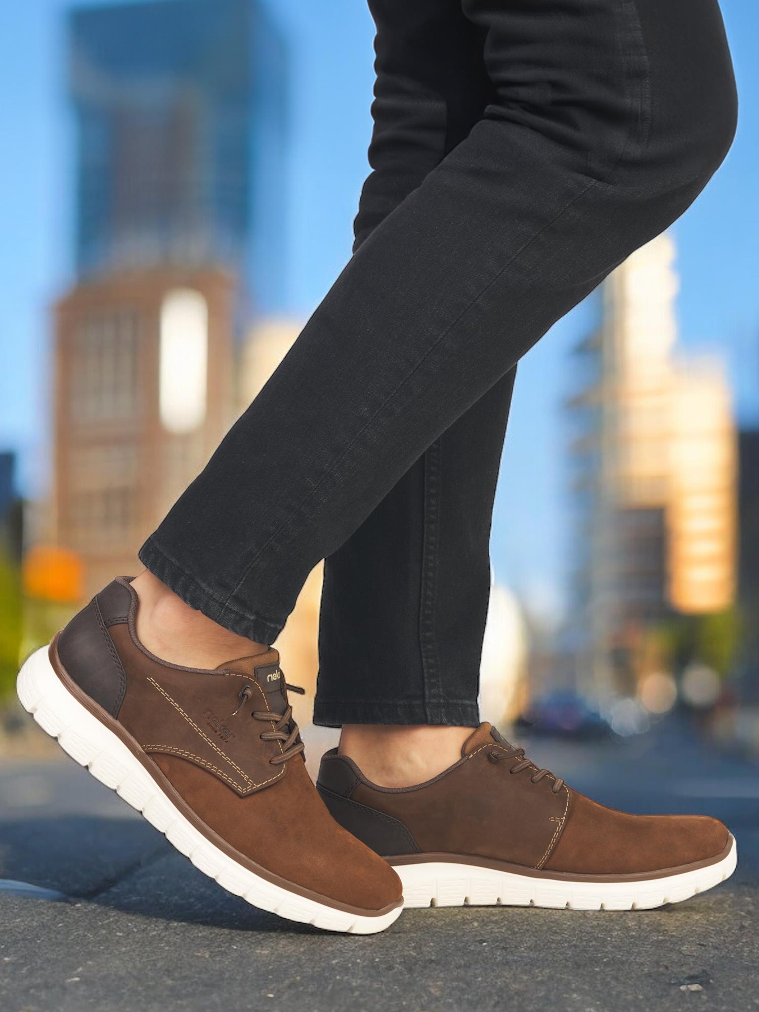 En person har på seg brune 07550 Rieker Antistress Walker-sko fra Rieker Comfort & Style med hvite såler og svarte bukser, og nyter overlegen komfort og et elegant utseende mens hun står i en bygate med uskarpe bygninger i bakgrunnen.