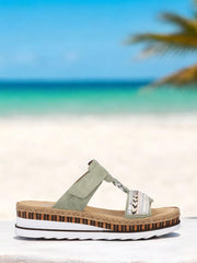 07518 Rieker Nature Soft-sandalen fra Rieker Comfort & Style har en lysegrønn stropp, vevde detaljer og en tykk platåsåle i kork og hvit, vist på hvitt med en uskarp strand- og havbakgrunn.