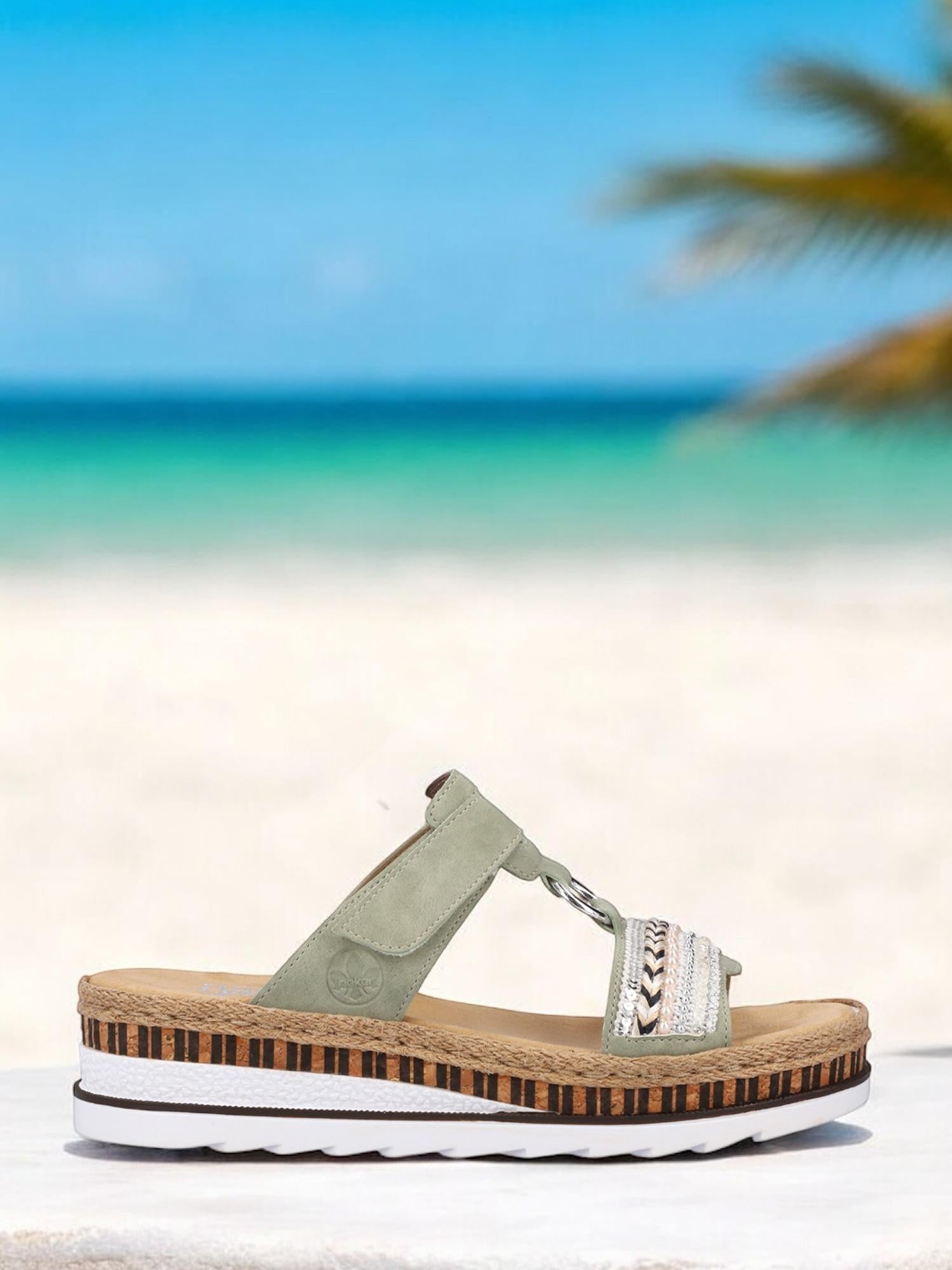 07518 Rieker Nature Soft-sandalen fra Rieker Comfort & Style har en lysegrønn stropp, vevde detaljer og en tykk platåsåle i kork og hvit, vist på hvitt med en uskarp strand- og havbakgrunn.