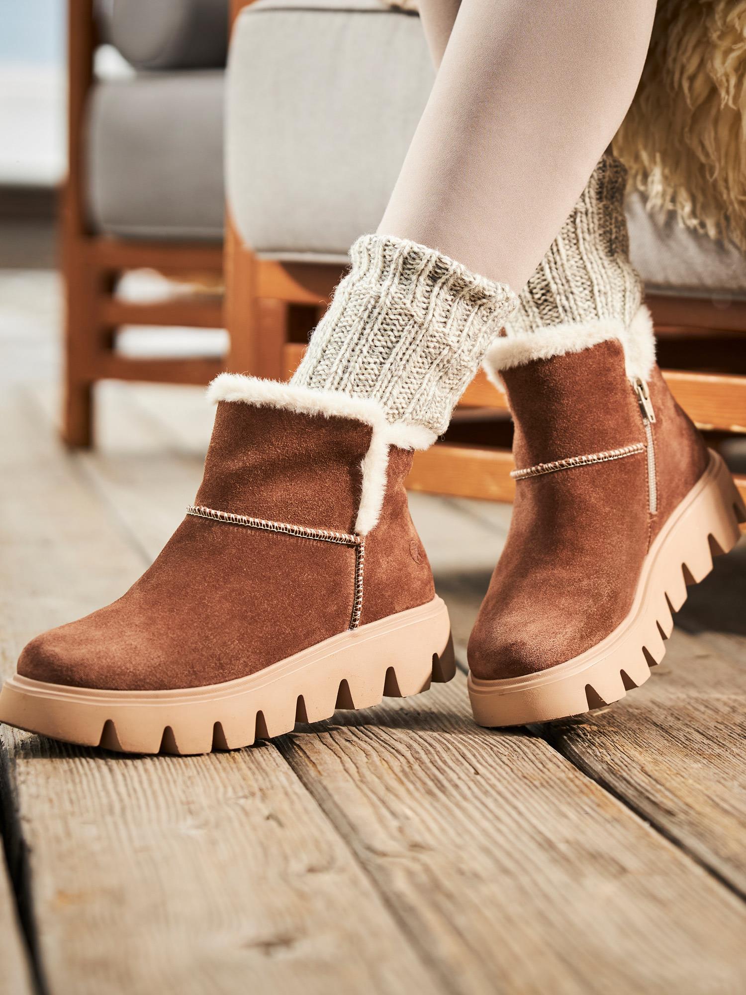 Nærbilde av en person iført Rieker Sport 05037 Rieker-Evolution Semsket ullfôret støvletter med hvit shearlingbesetning og chunky beige såler, stylet med kremfargede strikkede sokker, på et tregulv ved siden av en stol.