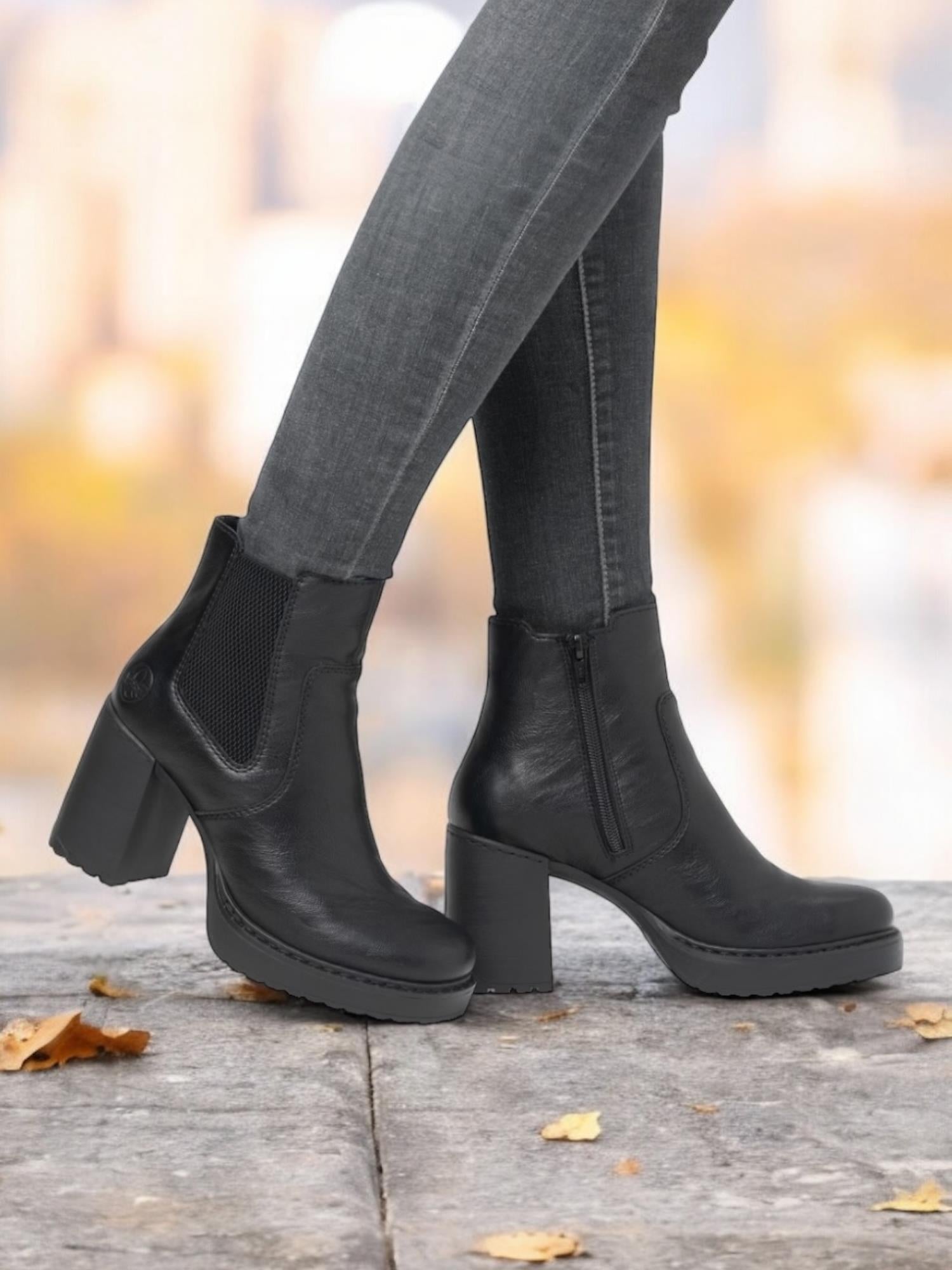 En person i mørke skinny jeans og svarte 04742 Rieker Heeled booties fra Rieker Comfort & Style står utendørs på en steinflate med spredte høstløv, og den uskarpe bakgrunnen understreker komfort og stil.