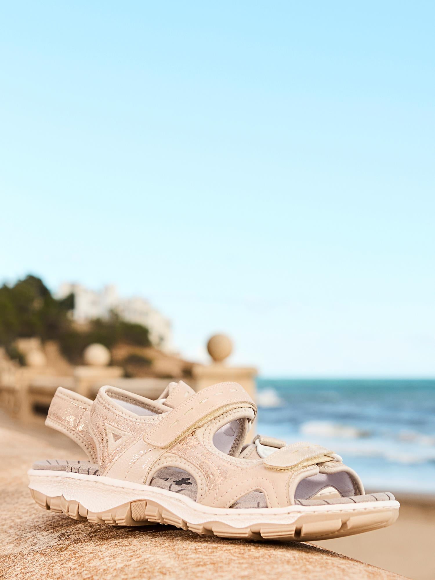 Et par 03024 Rieker Rose ComfortWave-sandaler fra Rieker Comfort & Style, med hvite såler, hviler på en steinhylle ved havet med et mykt uskarpt bilde av havet, himmelen, fjerne bygninger og grøntområder i bakgrunnen.