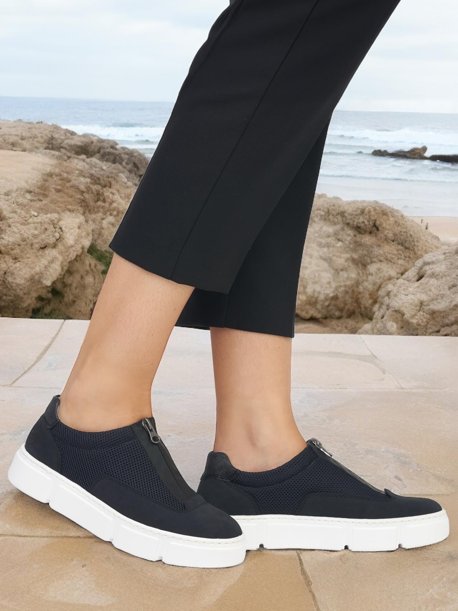 En person som har på seg svarte Rieker Comfort & Style 07577 Summer Zip-on-sneakers med hvite såler og svarte bukser står på en steinete overflate ved et steinete terreng, med havet i bakgrunnen.