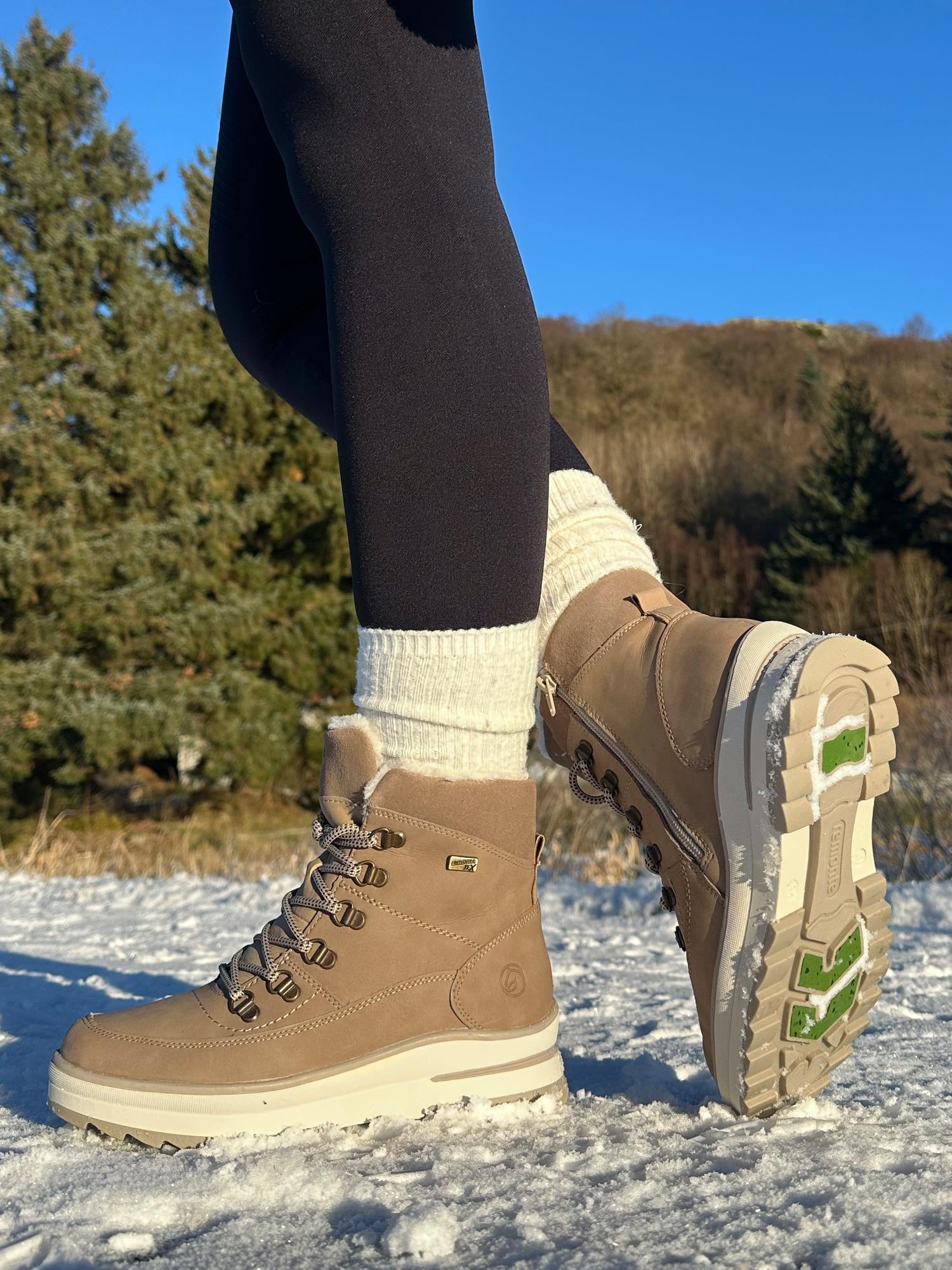 En person iført Remonte 05022 Fiber-Grip tex-membran skinn ullfôr beige støvler, hvite sokker og svarte leggings står på snødekt mark med trær og blå himmel i bakgrunnen.