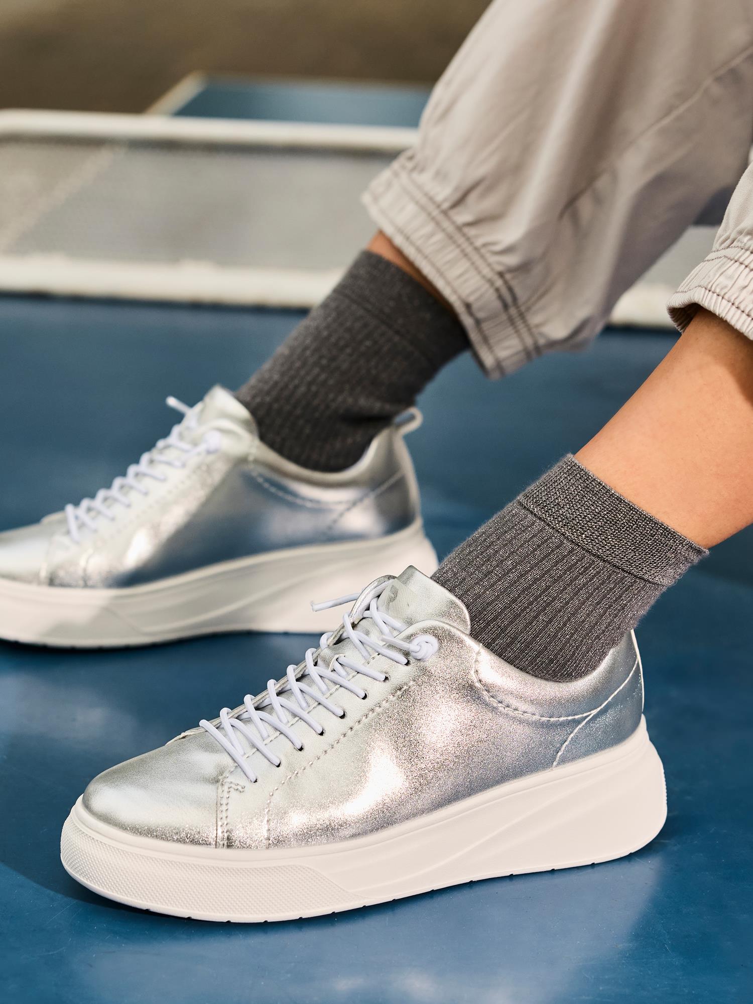 Et nærbilde viser en person som har på seg Rieker Sport 03545 ShineWalk sølvfargede skinnsneakers med hvite lisser og tykke hvite såler, kombinert med grå ribbede sokker og lyse bukser med mansjetter, stående på en blå overflate.