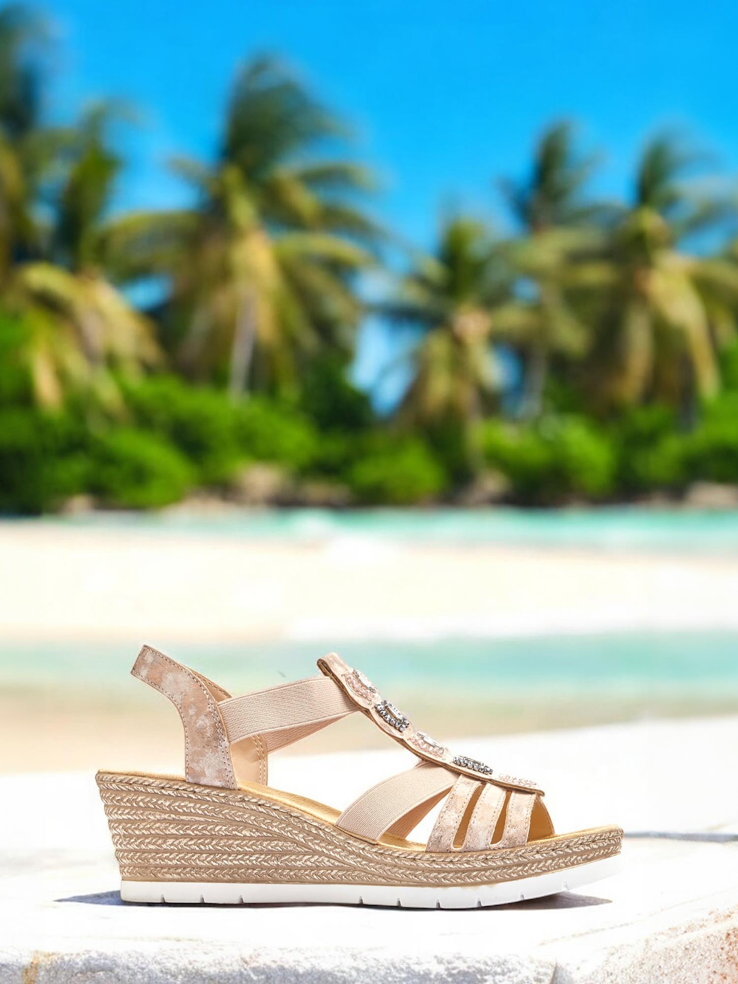 07636 Rieker Zahara Stone-sandalen fra Rieker Comfort & Style, med beige stropper og dekorative perler, vises utendørs på en hvit avsats med palmer, sandstrand og turkis hav i bakgrunnen.