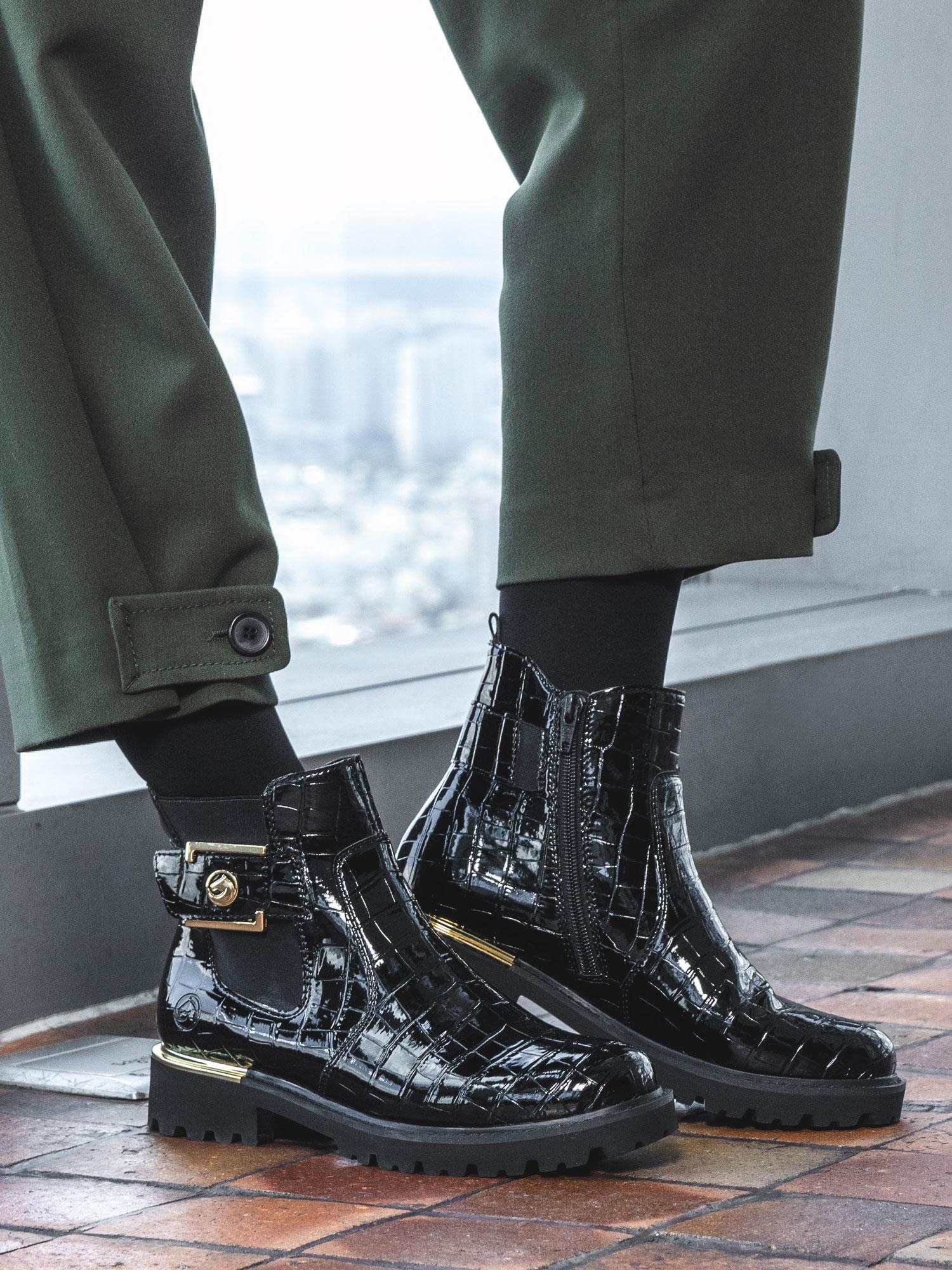 En person i mørkegrønne bukser og Remontes 05024 Gold Croc Boot med tykke såler, gulldetaljer og glidelåser på siden står på et flislagt gulv ved et vindu med utsikt over byen.