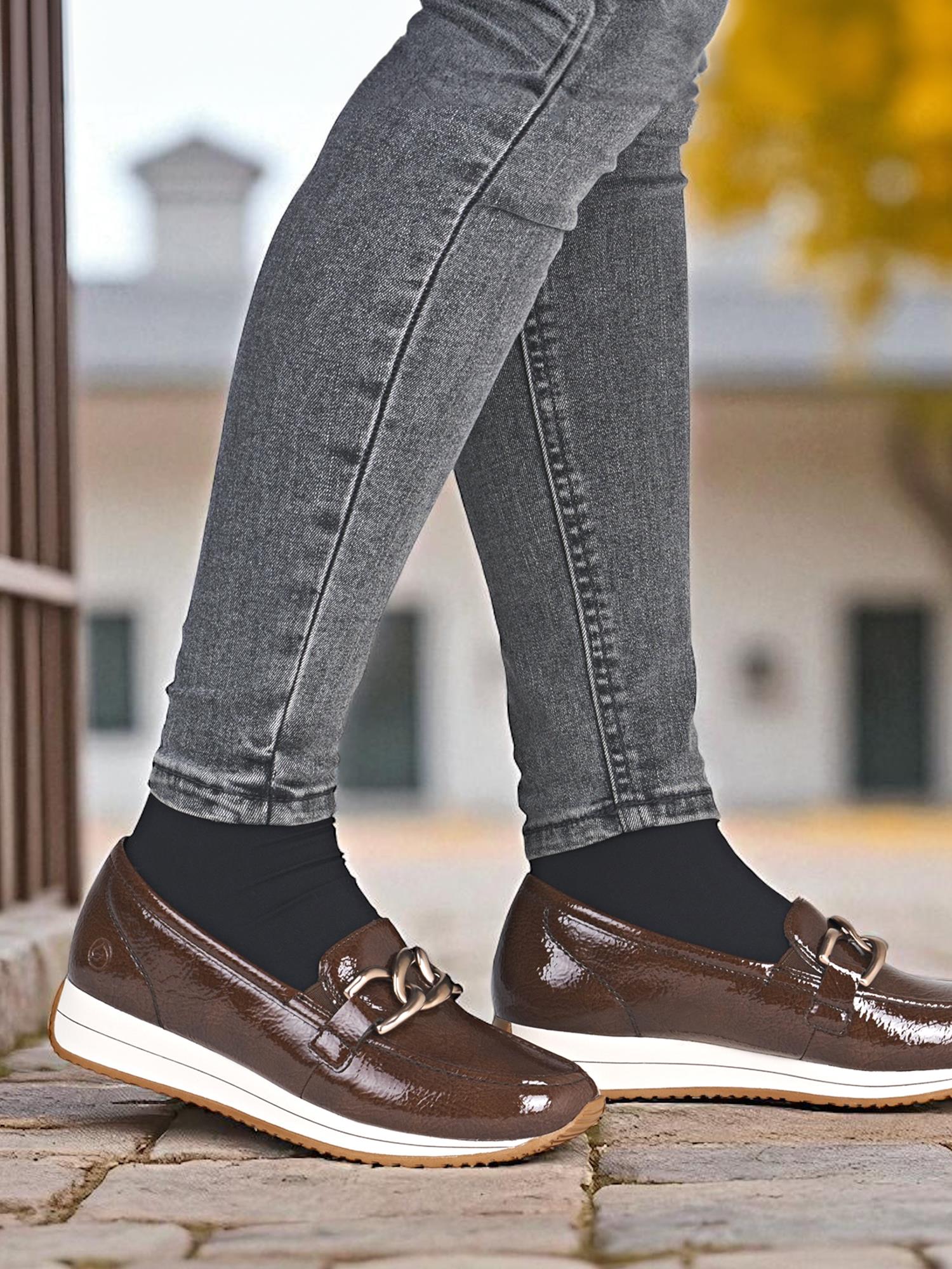 En person står utendørs på brostein iført grå skinny jeans, svarte sokker og 04547 Remonte Liona Mokkasin fra Rieker Comfort & Style, med tykke, hvite såler og metallspenne.