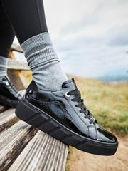 En person iført 04543 Rieker-Evolution Patent Leather Platform Sneaker fra Rieker Sport, med grå sokker og svarte leggings, sitter på en trebenk utendørs med gress og en overskyet himmel i bakgrunnen.