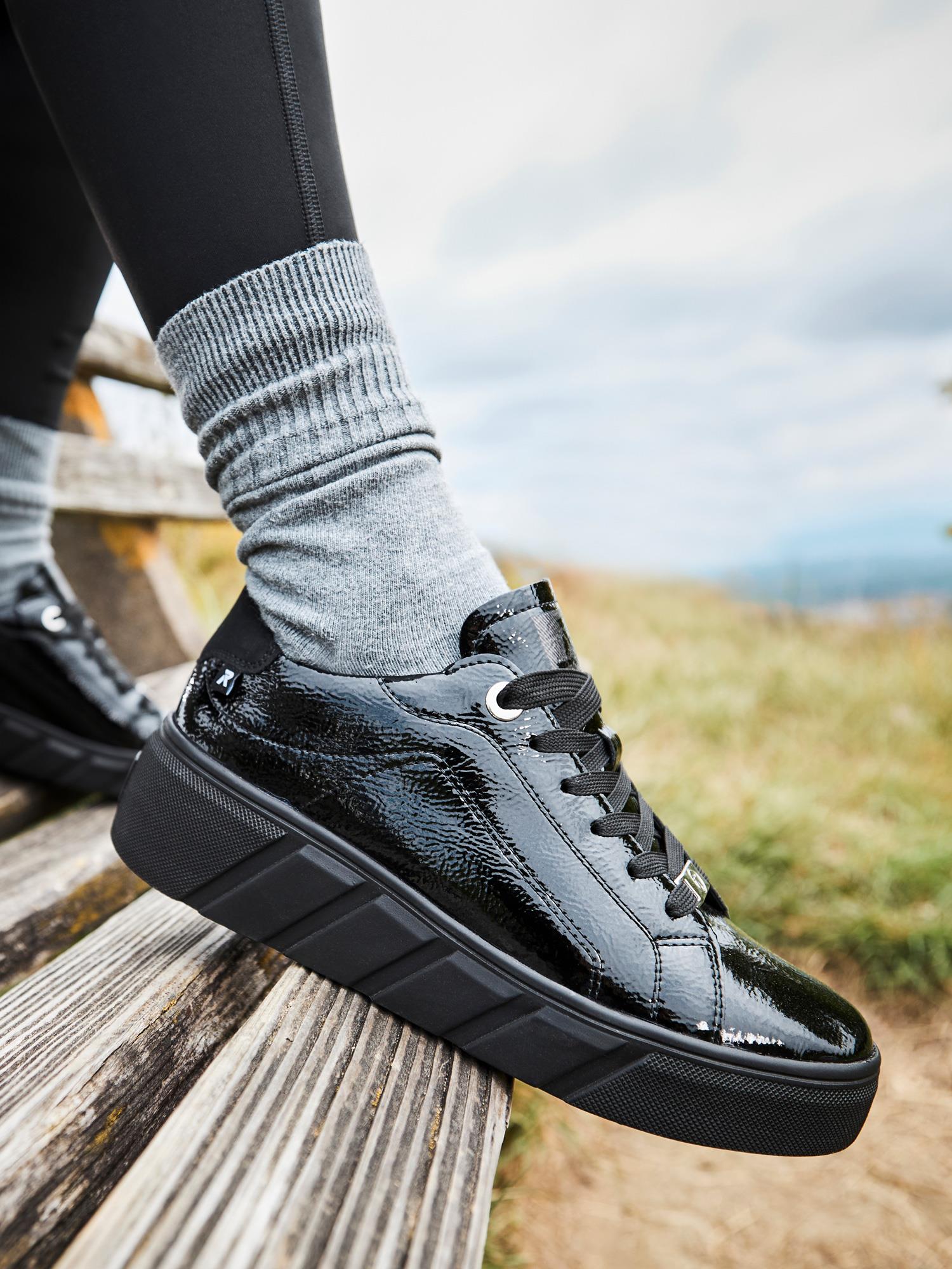 En person iført 04543 Rieker-Evolution Patent Leather Platform Sneaker fra Rieker Sport, med grå sokker og svarte leggings, sitter på en trebenk utendørs med gress og en overskyet himmel i bakgrunnen.