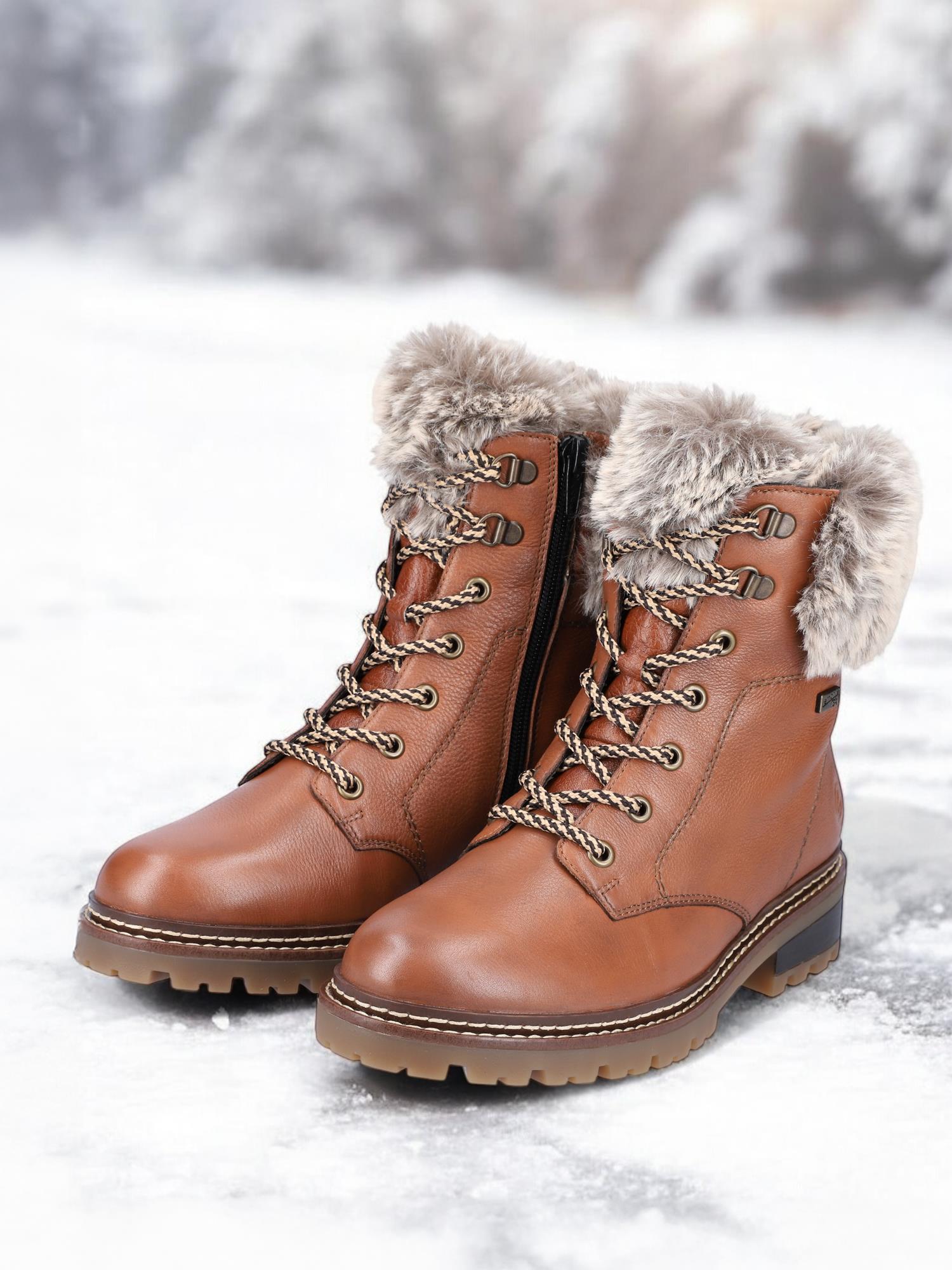 Et par 04801 Remonte vintersko i brunt skinn med pelsfôr og såler med fibergrip er vist på snø, mot en uskarp skogsbakgrunn.