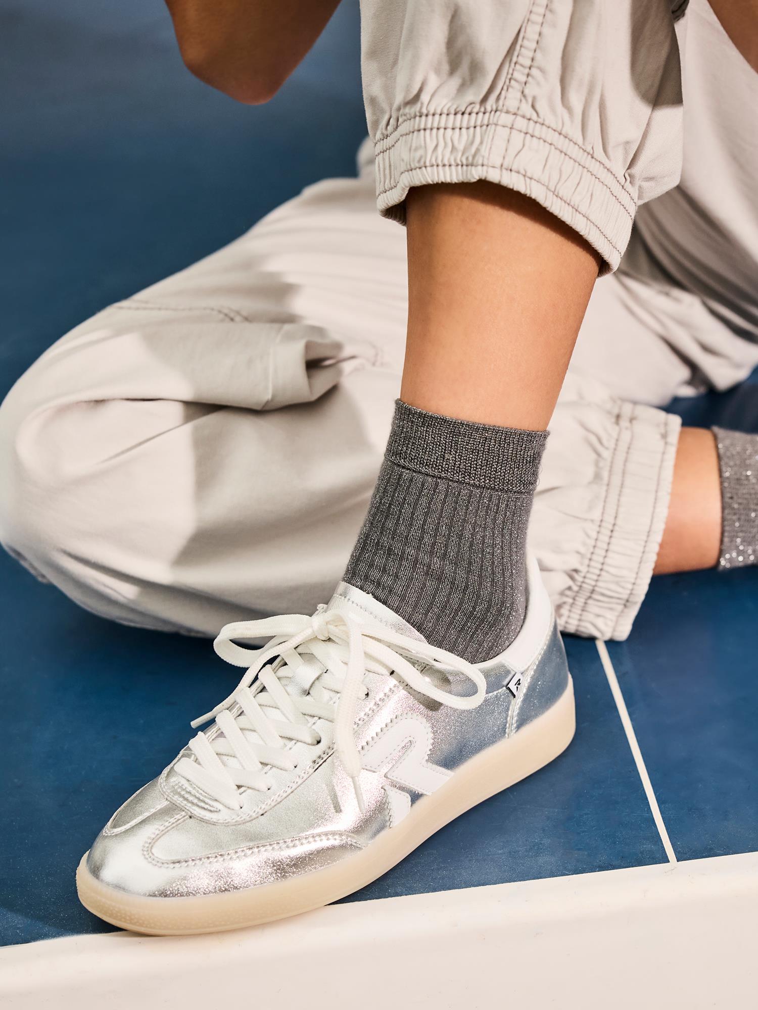 En person i lysegrå bukser med mansjetter og ribbede grå sokker sitter på et blått gulv og viser frem 03524 Rieker San Diego Leather Sneaker Silver fra Rieker Sport med hvite lisser.