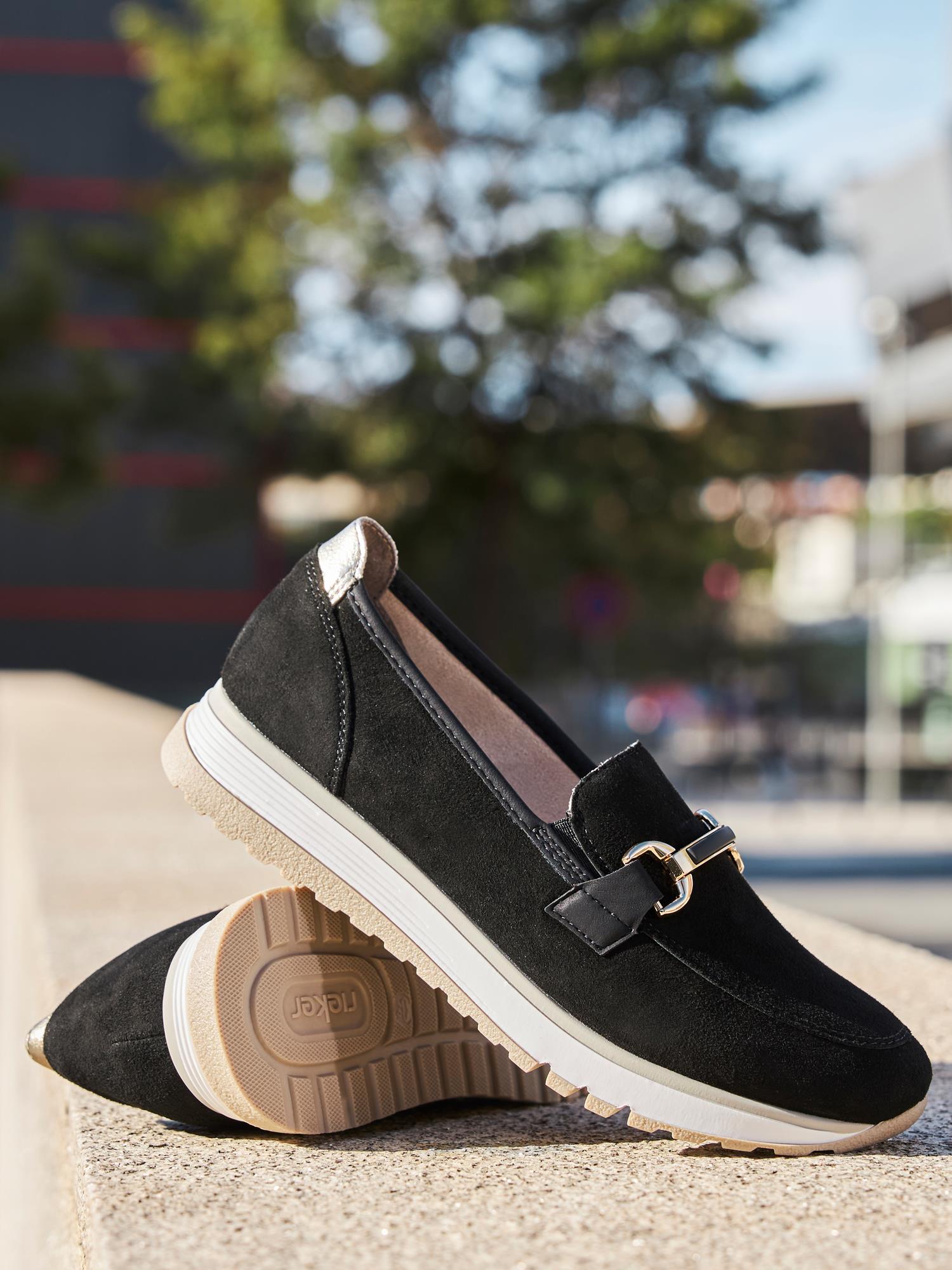 02959 Rieker Soft Elegance Leather Black loafers fra Rieker Comfort & Style, med hvit såle og gullspenne, vises utendørs mot et urbant bakteppe - en uanstrengt blanding av komfort og eleganse.