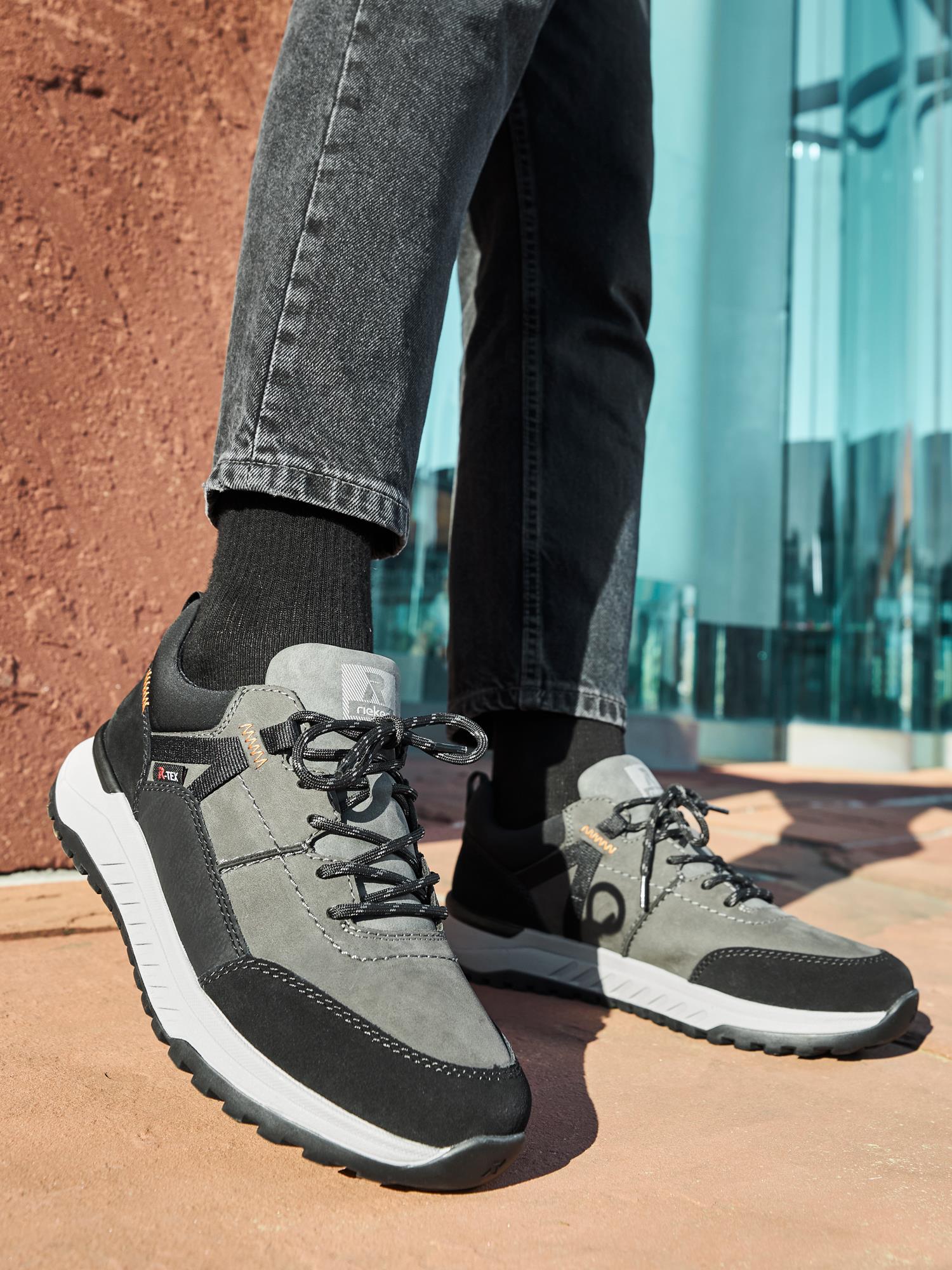 En person i grå og svarte Rieker Sport 04504 Evolution Peak Sneaker vanntette sko med hvite såler og svarte sokker står på en solrik utendørs overflate ved en teksturert rød vegg og reflekterende glasspaneler.