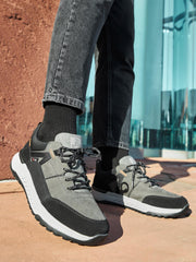 En person i grå og svarte Rieker Sport 04504 Evolution Peak Sneaker vanntette sko med hvite såler og svarte sokker står på en solrik utendørs overflate ved en teksturert rød vegg og reflekterende glasspaneler.