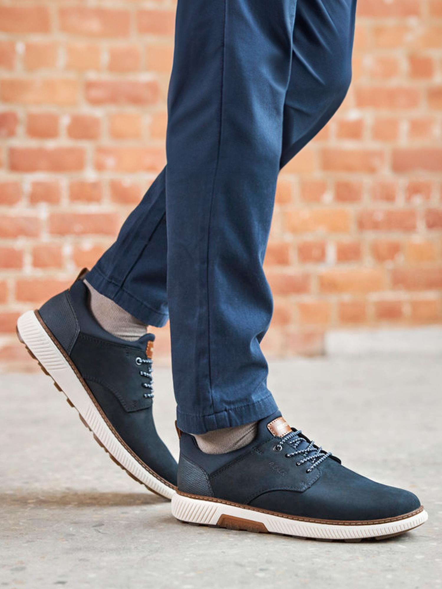 En person i marineblå bukser og mørkeblå 05406 Rieker Softfoam Sneakers fra Rieker Comfort & Style, med hvite såler og Antistress-teknologi, står på et grått betonggulv foran en murvegg.