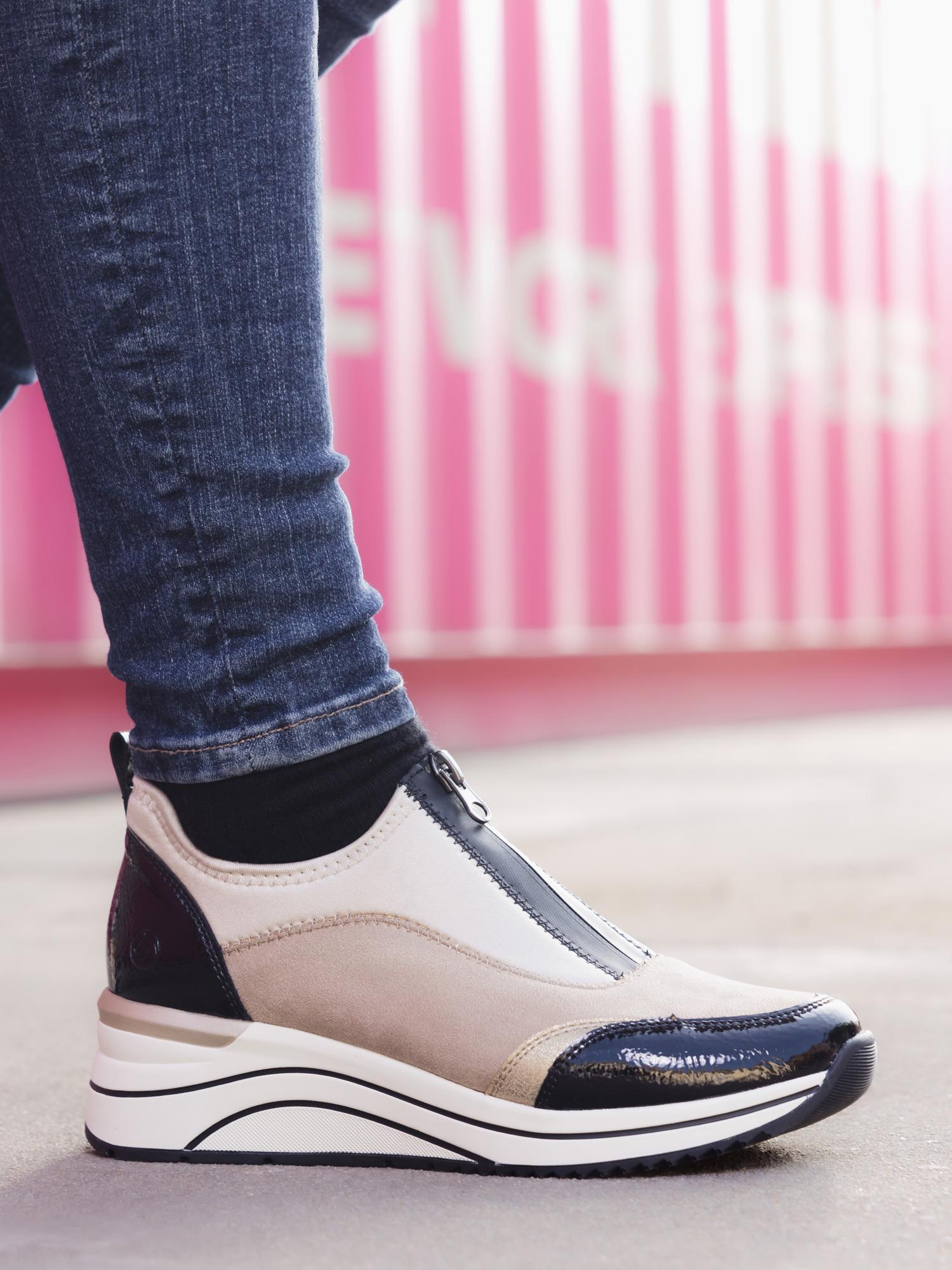 Iført skinny blå jeans og en svart sokk viser en person frem Remonte 04961 High Soft Multi Sneaker Beige – en beige og svart sneaker med glidelås og en tykk hvit og svart såle – stående på fortauet med en uskarp rosa og hvit bakgrunn.
