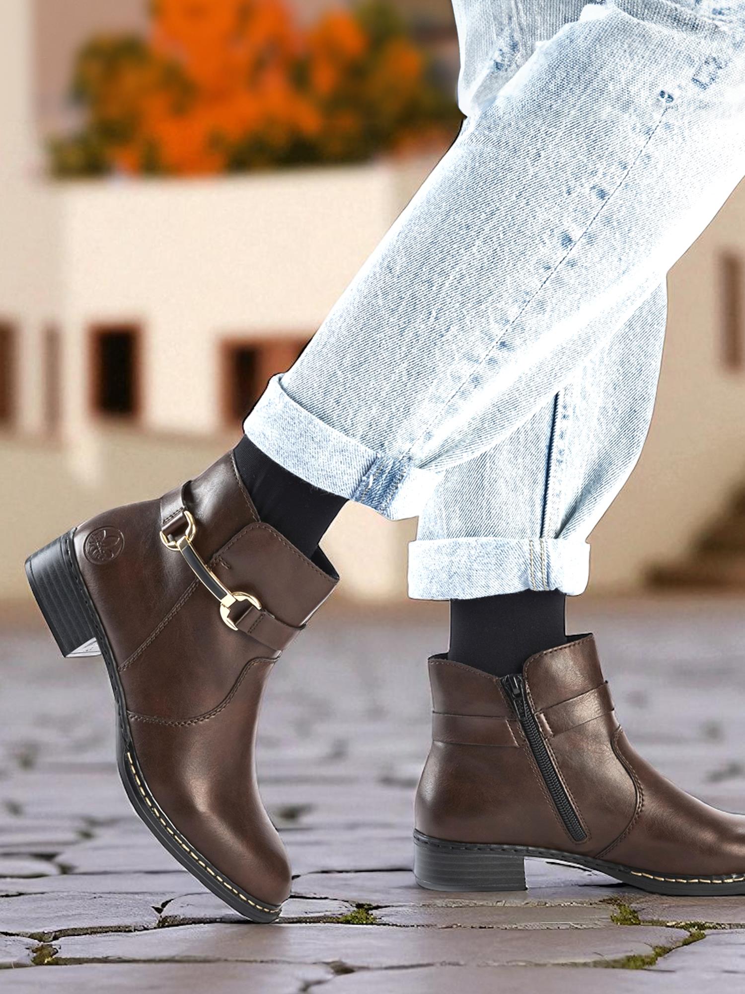 En person i lyse, opprullede jeans og 04314 Rieker Nalia Ankelstøvlett fra Rieker Comfort & Style står på en brosteinsbelagt gate, med en uskarp bygning og høstløv i bakgrunnen.