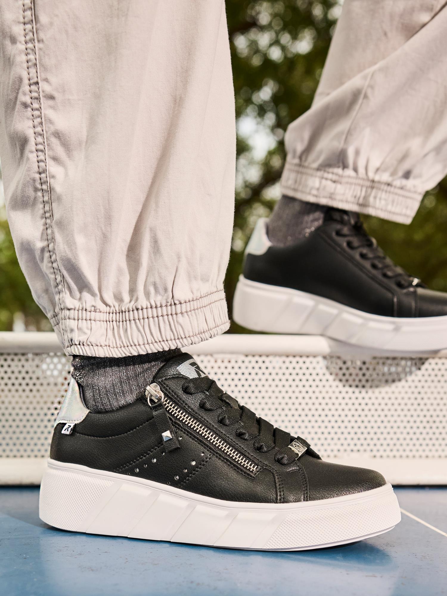 En person har på seg Rieker Sport 03558 Rieker Active Platform Leather-sneakers med hvite såler og sølvfargede glidelåser, pluss grå sokker og lyse bukser med mansjetter, stående utendørs på en blå og hvit overflate.