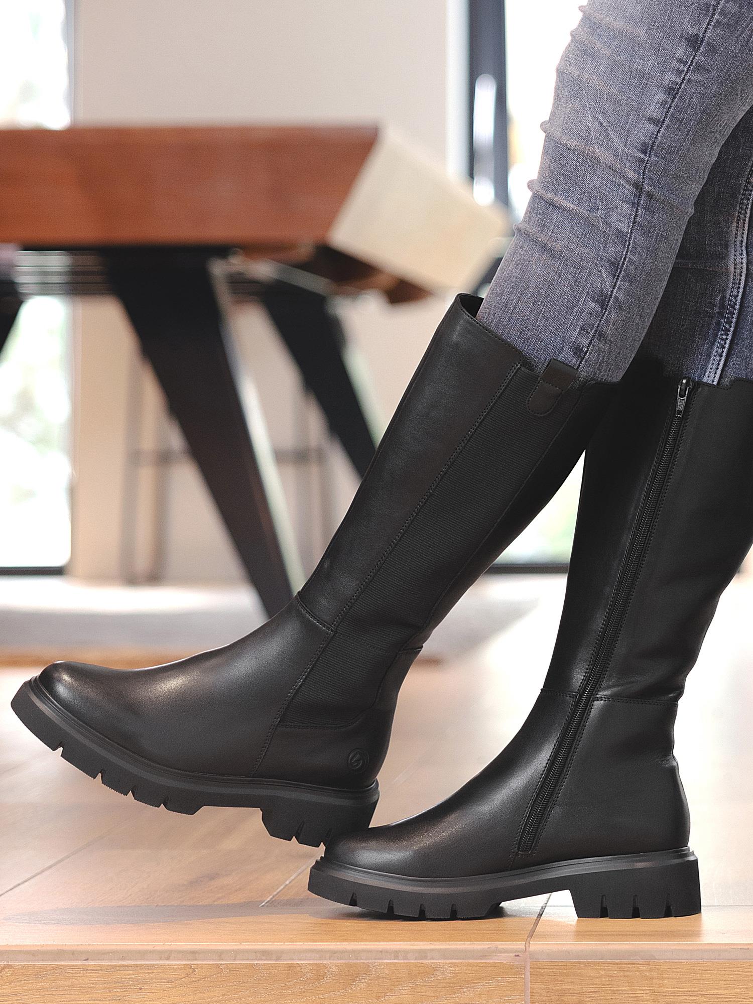 En person står innendørs på et tregulv, iført blå jeans og svarte, knehøye 04566 Remonte Celina Leather Boots fra Remonte, med tykke såler, glidelåser og skinnmateriale.