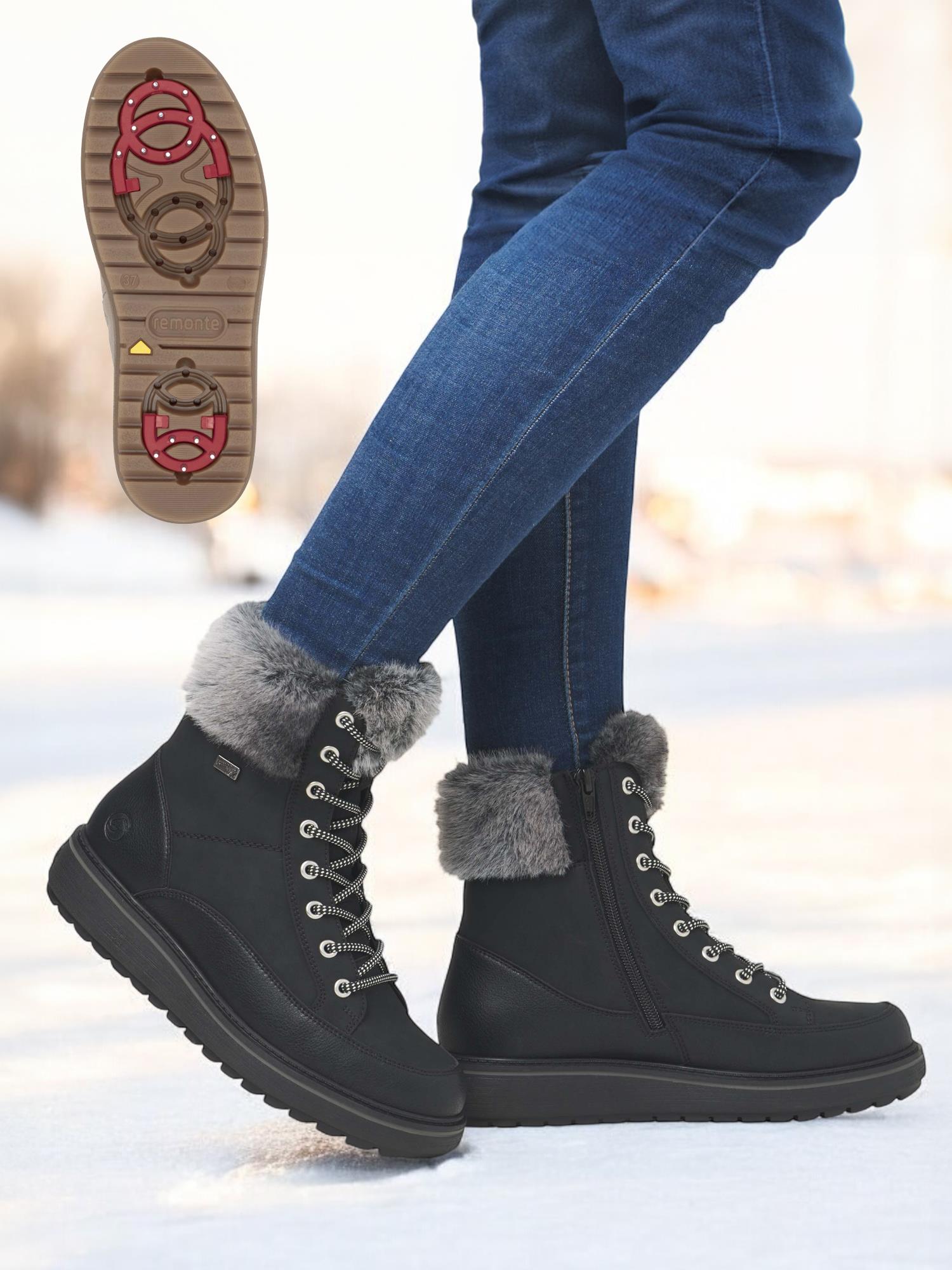 En person i blå jeans har på seg Remonte 05006 Premium leather tex sorte ullfôret med brodder vinterstøvler med grå fuskepels på snø. Et innfelt viser sålens røde og brune mønster for ekstra godt grep.