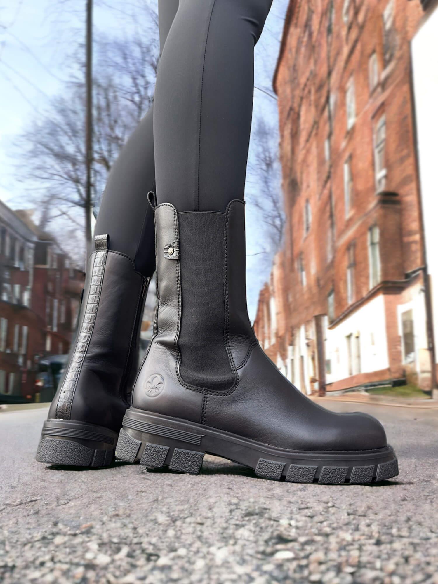En person står i en bygate i svarte leggings og Rieker Comfort & Style 03046 Classic chelsea varmfôret vinterstøvler i skinn, belyst fra en lav vinkel for å vise det stilige utseendet og det varmende fôret for ekstra komfort. I bakgrunnen ser vi murbygninger og trær.