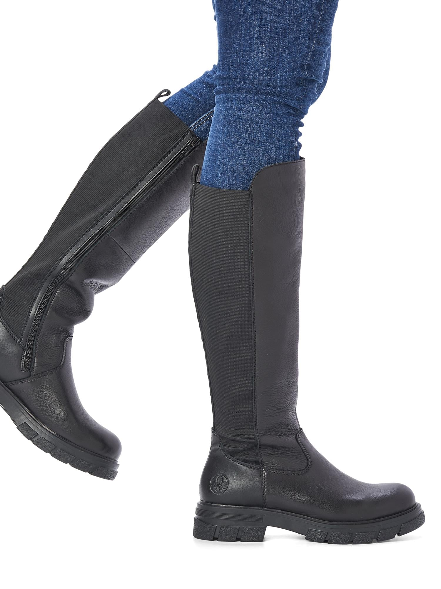 En person iført blå jeans og 05368 Rieker High Elastic Trend Boots fra Rieker Comfort & Style trer frem mot en hvit bakgrunn.