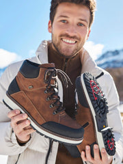 En smilende mann i hvit jakke holder opp et par 04386 Rieker Mandel Varmfòret med Brodder vintersko fra Rieker Sport, med svarte såler, utendørs i snøen og fjellene.