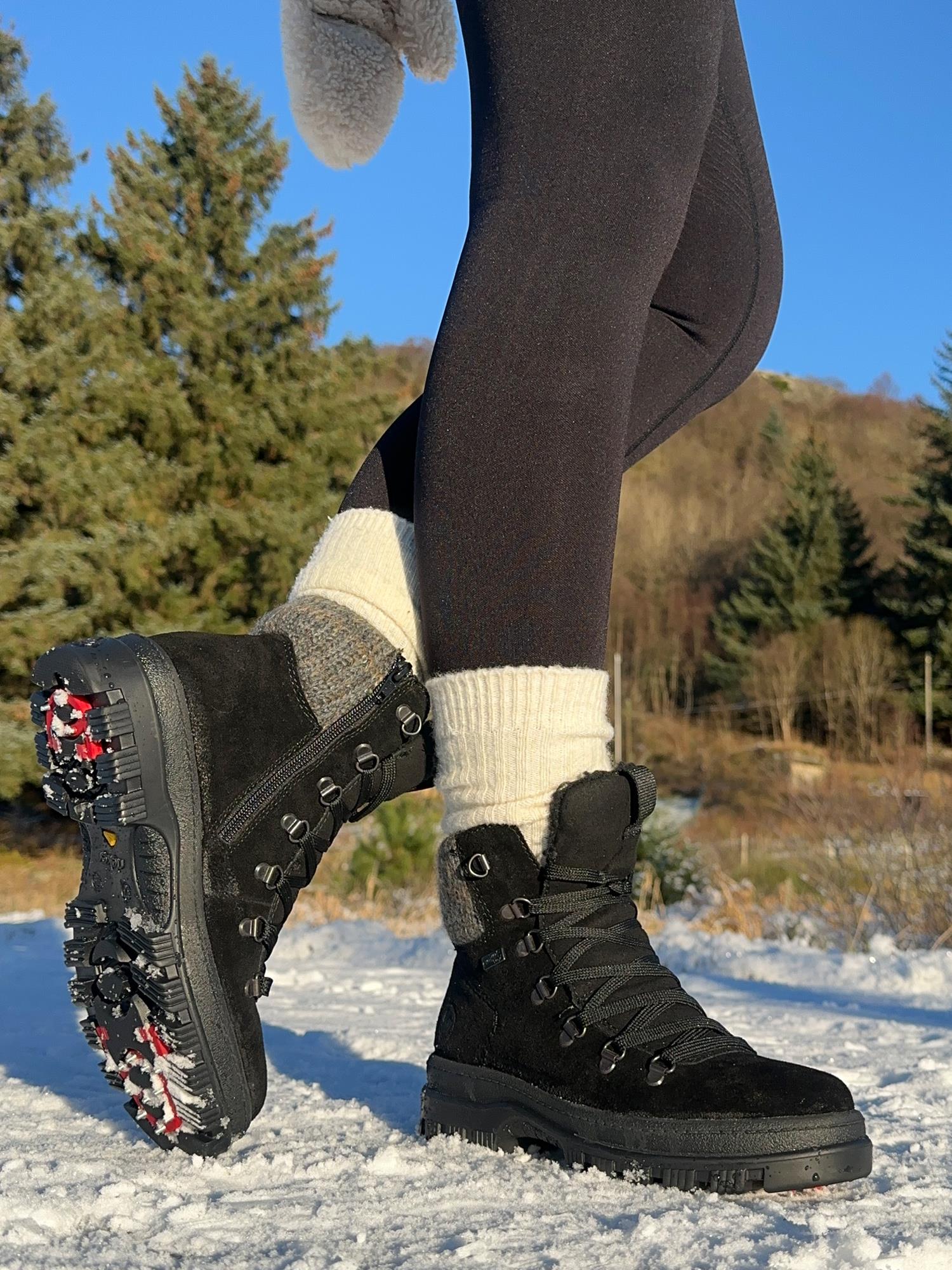 En person i svart 05364 Rieker Varmfòret vintersko med brodder fra Rieker Comfort & Style, tykke ullsokker og svarte leggings står på snødekt mark med røde pigger og snø på sålene. I bakgrunnen skimtes trær og åser.
