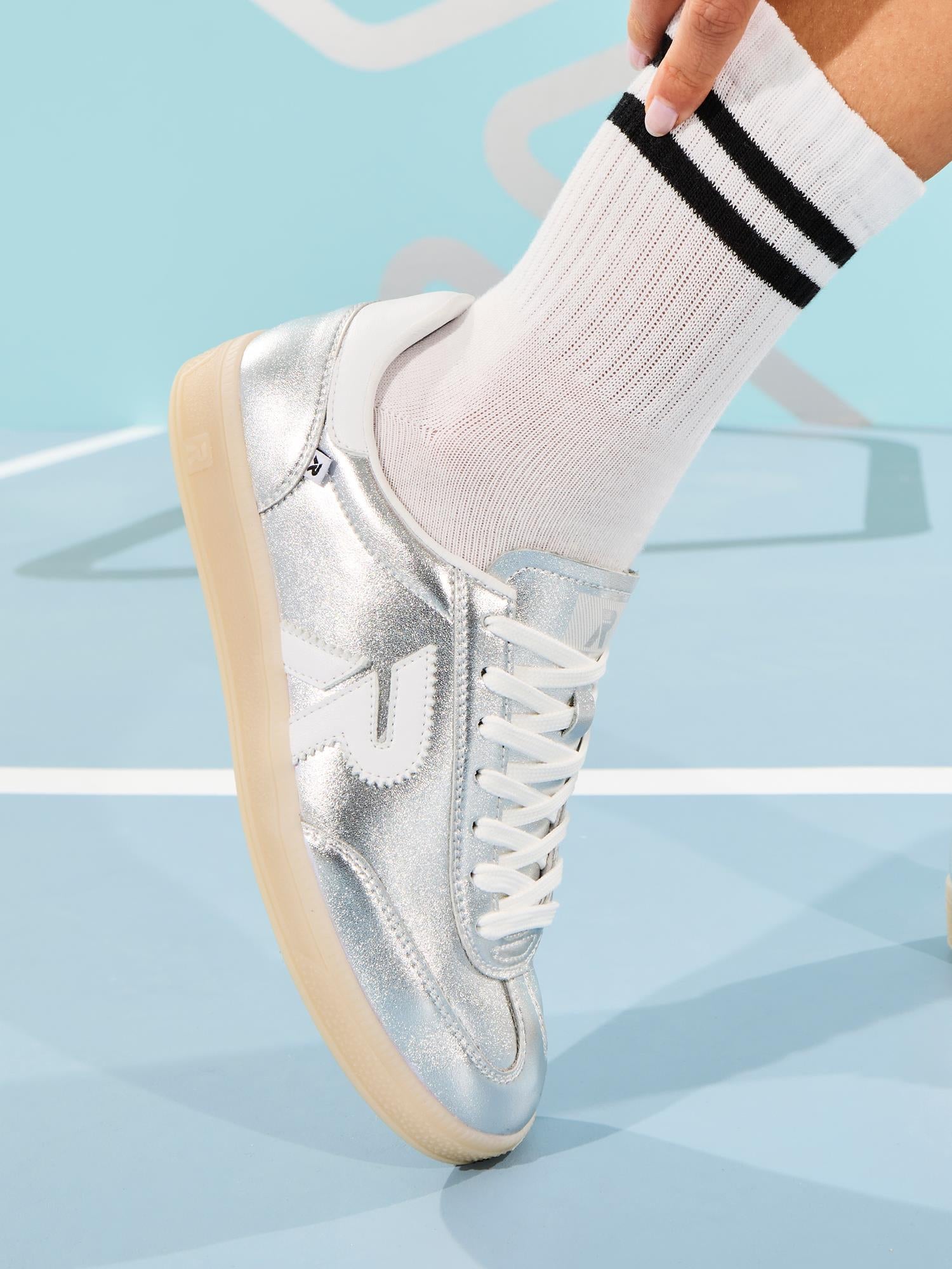 Noen står på en lyseblå, geometrisk mønstret overflate iført hvite sokker med svarte striper og Rieker Sport 03524 Rieker San Diego Leather Sneaker Silver med hvite lisser og en hvit logo.