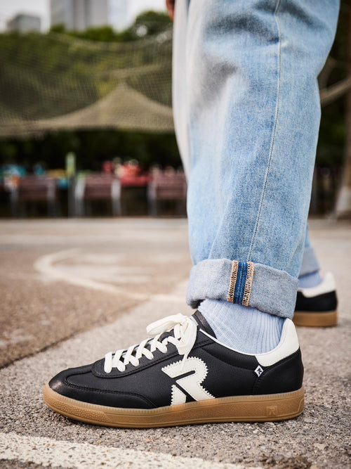 En person iført lyseblå jeans med mansjett og svarte Rieker Sport 03332 San Diego Leather Sneakers står på en utendørsbane, med trær, benker og et nett synlig i den uskarpe bakgrunnen.