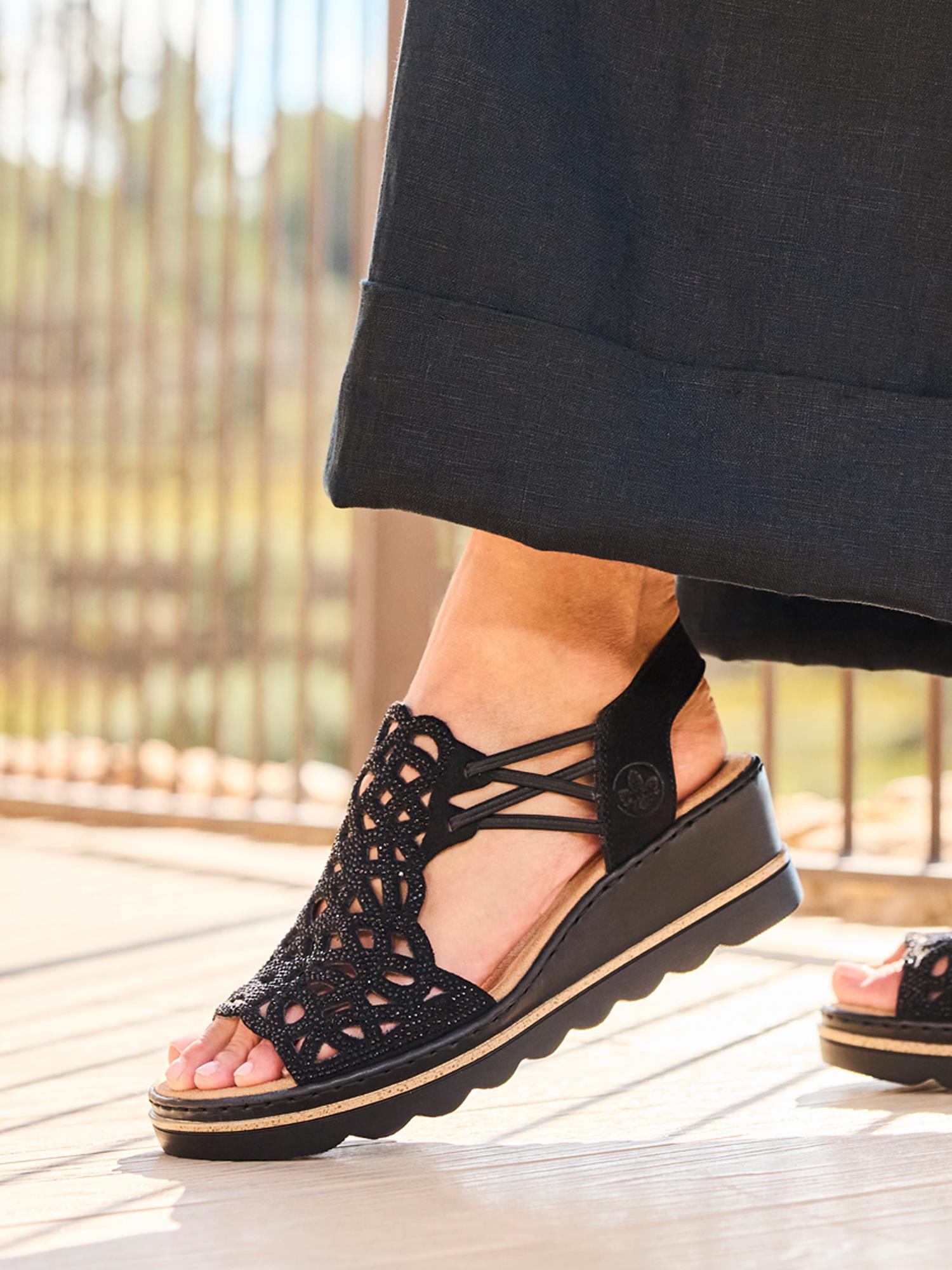 En person i langt svart skjørt og 03003 Rieker Sorella Sandal fra Rieker Comfort & Style-en komfortabel damesandal med intrikate cut-out-mønstre-står på et solbelyst tregulv.