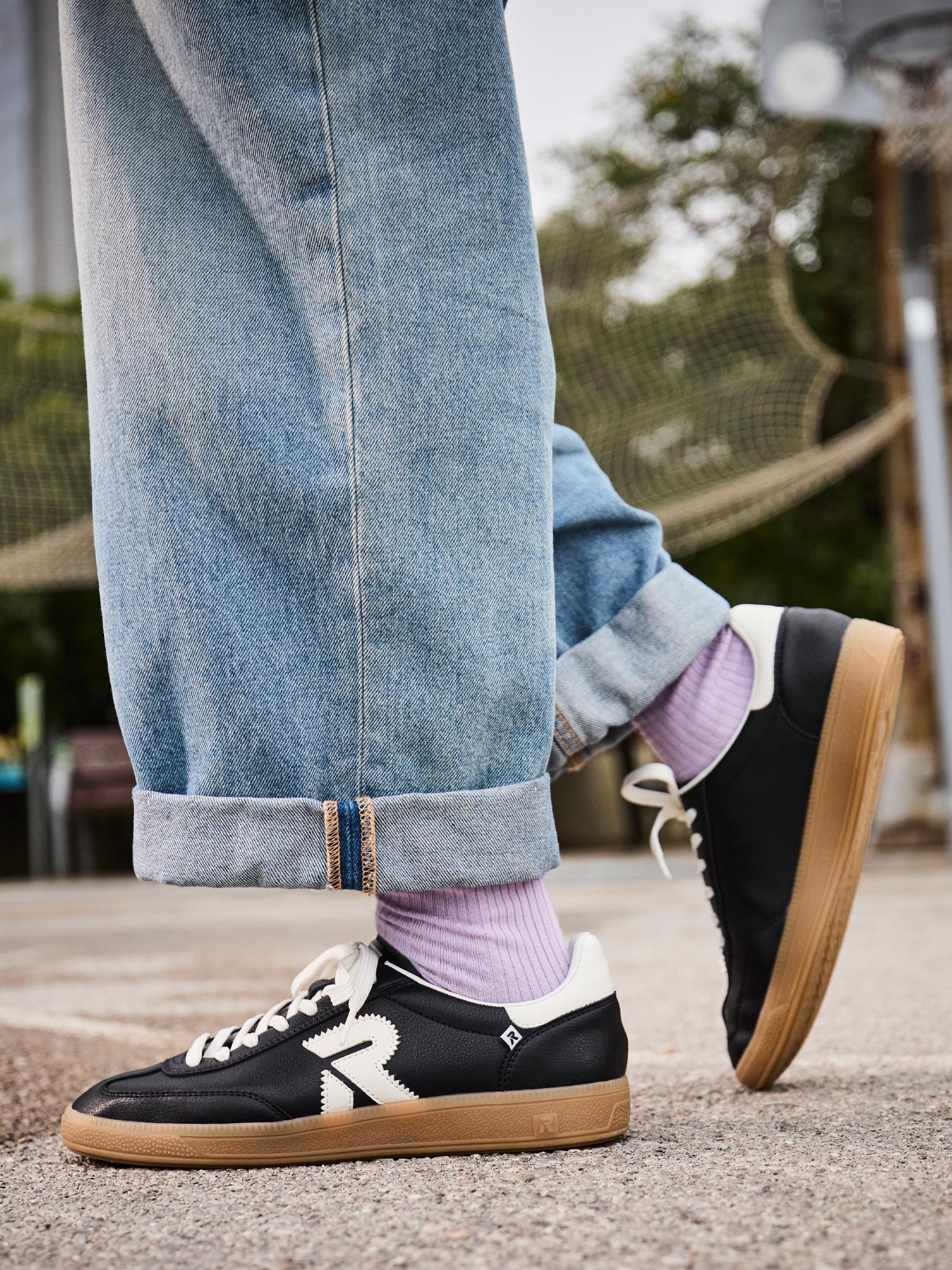 En person står utendørs på en basketballbane iført løse blå jeans med mansjetter, lyselilla sokker og Rieker Sport 03470 San Diego skinnsko til dame, svart med hvite detaljer og gummisåler.