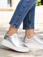 En person i skinny blå jeans og Rieker Sport 03545 ShineWalk sølvfargede skinnsneakers med tykke hvite såler står utendørs på et solfylt fortau.