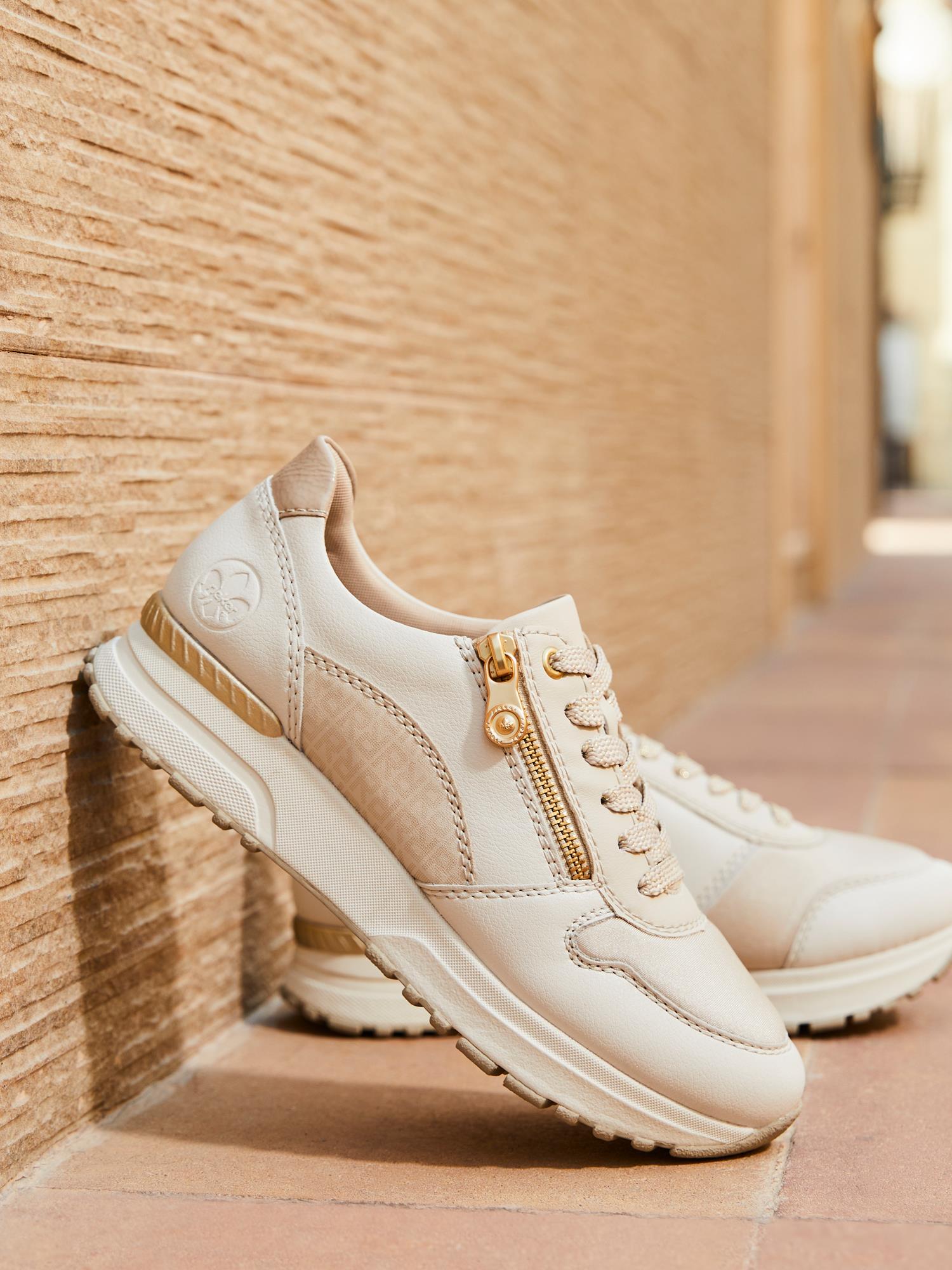 Rieker Comfort & Style 07580 Beige Leather Zip Sneaker har gulldetaljer, glidelåser på siden, Antistress-teknologi, snørebånd, logo på hælen og en utskiftbar innersåle for ekstra komfort, som vises mot en teksturert vegg.