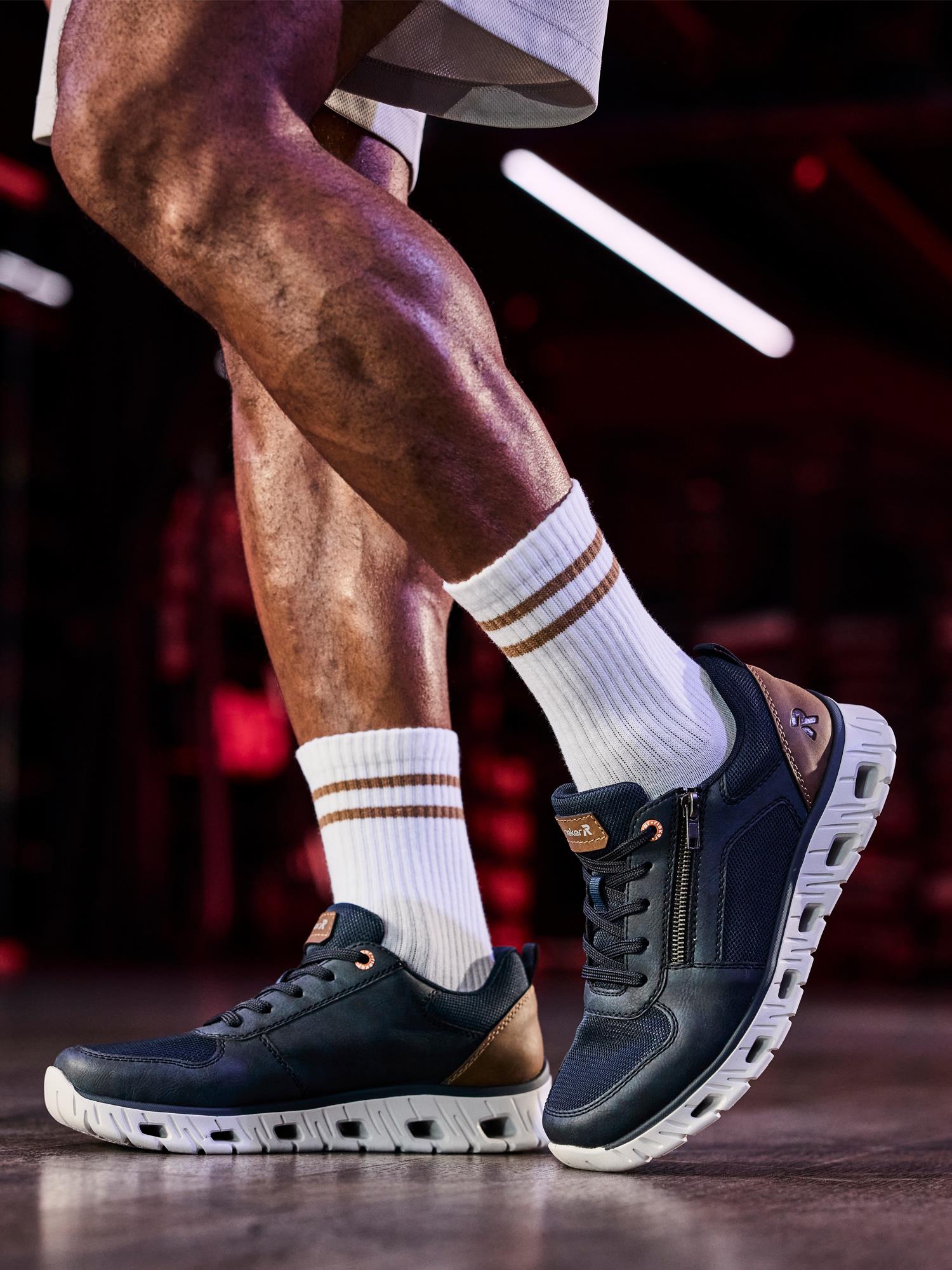 Nærbilde av en person som bruker Rieker Sport 02877 AirBounce Sneakers Navy i syntetisk skinn med hvite såler og hvite ankelsokker, innendørs under rosa-rød belysning som fremhever det komfortable fottøyet.