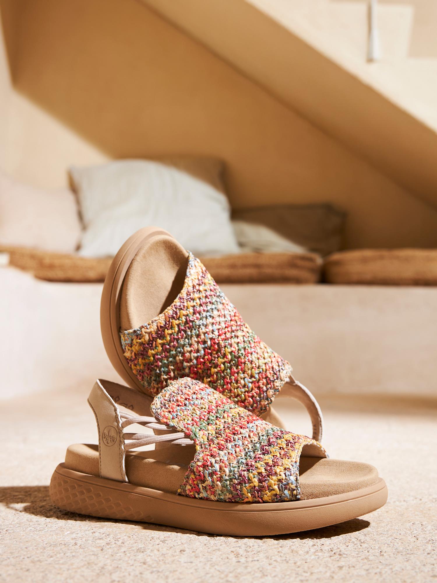 Et par 03341 Rieker Viala Sandals fra Rieker Comfort & Style - flerfarget med solbrun såle - hviler på et solbelyst innendørsgulv, og gir fargerik komfort mot uskarpe puter og et koselig bakteppe.