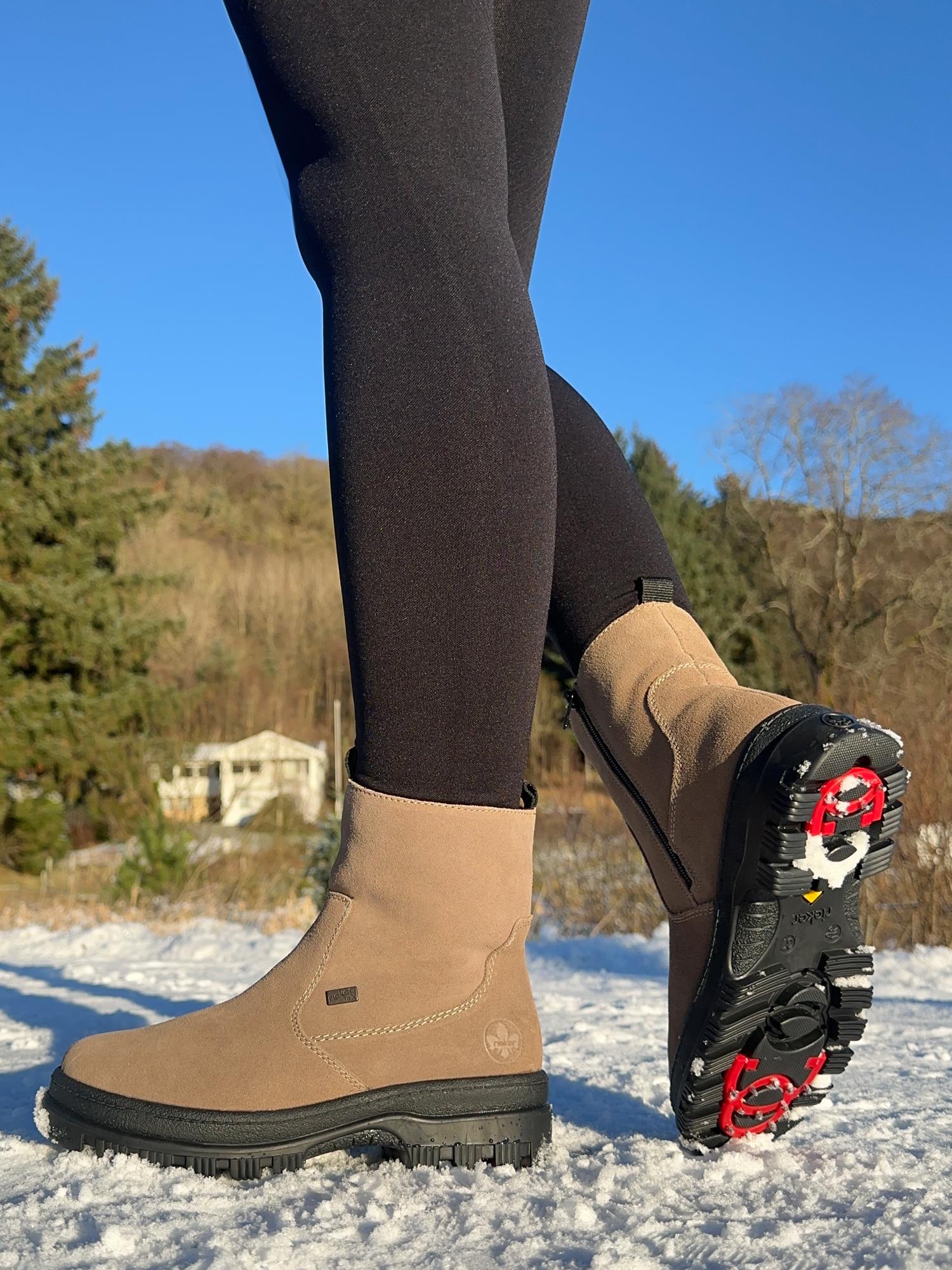 Iført 05039 Rieker Leather Tex varmforet med brodder fra Rieker Comfort & Style står en person på snø foran uskarpe eviggrønne planter og et hus under en klar himmel.