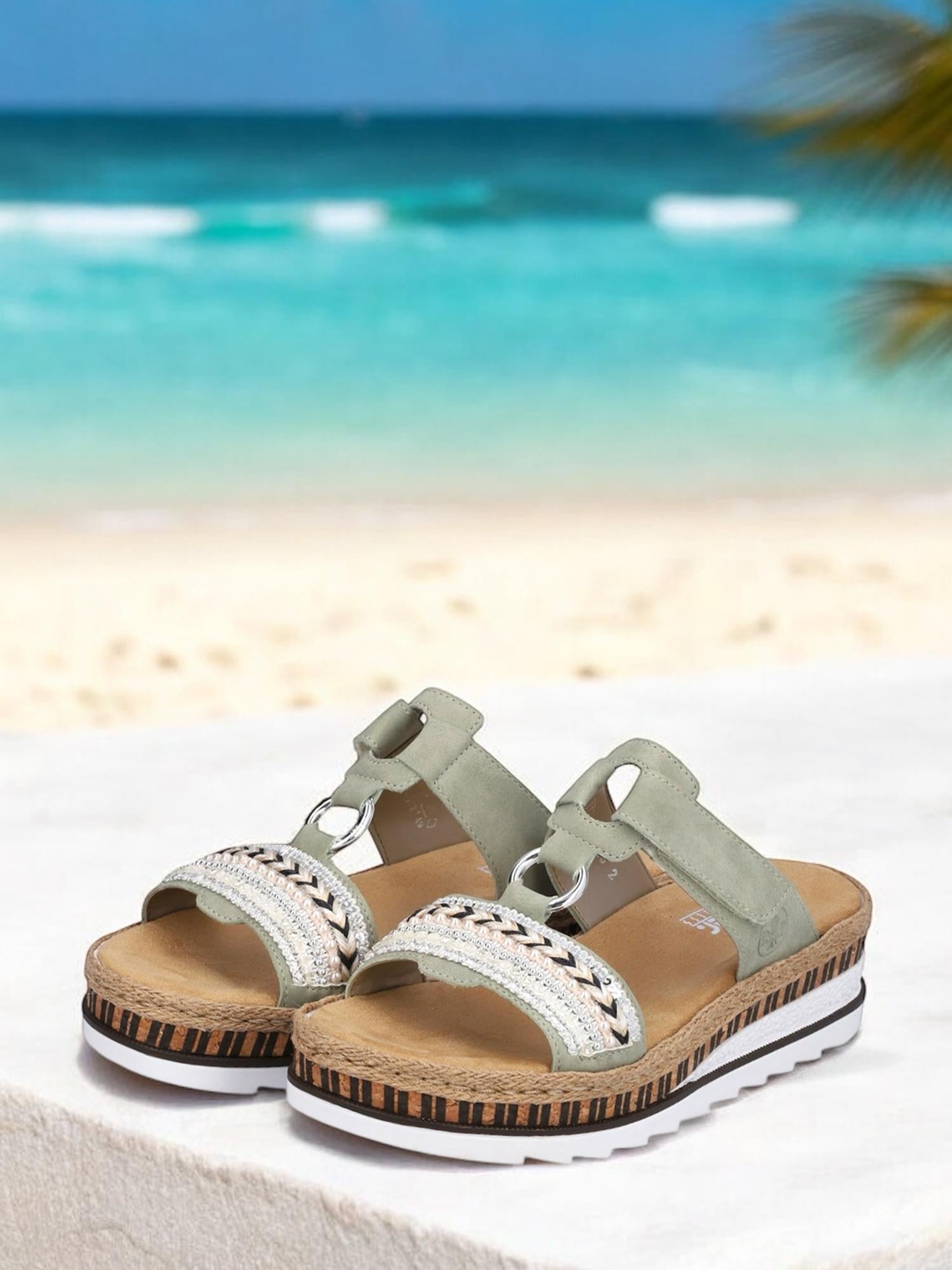 Et par 07518 Rieker Nature Soft Sandaler fra Rieker Comfort & Style med lysegrønne stropper, sølvdetaljer og korksåler vises på en hvit overflate mot en tropisk strand og turkis hav for ekstra komfort.