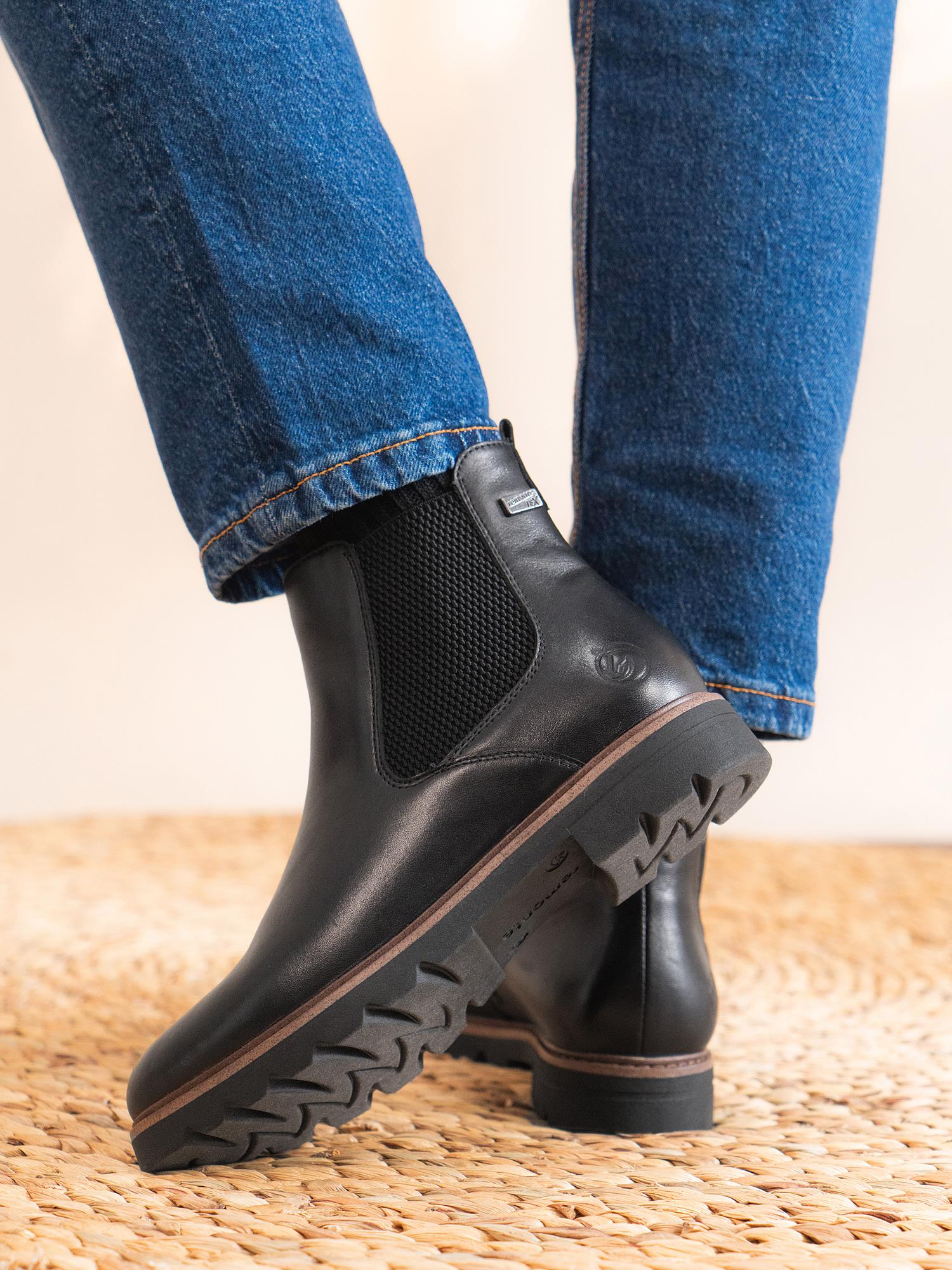 En person iført blå jeans og Remonte 04571 Torino ankelstøvler i skinn med tykke, robuste såler står på en vevd matte.