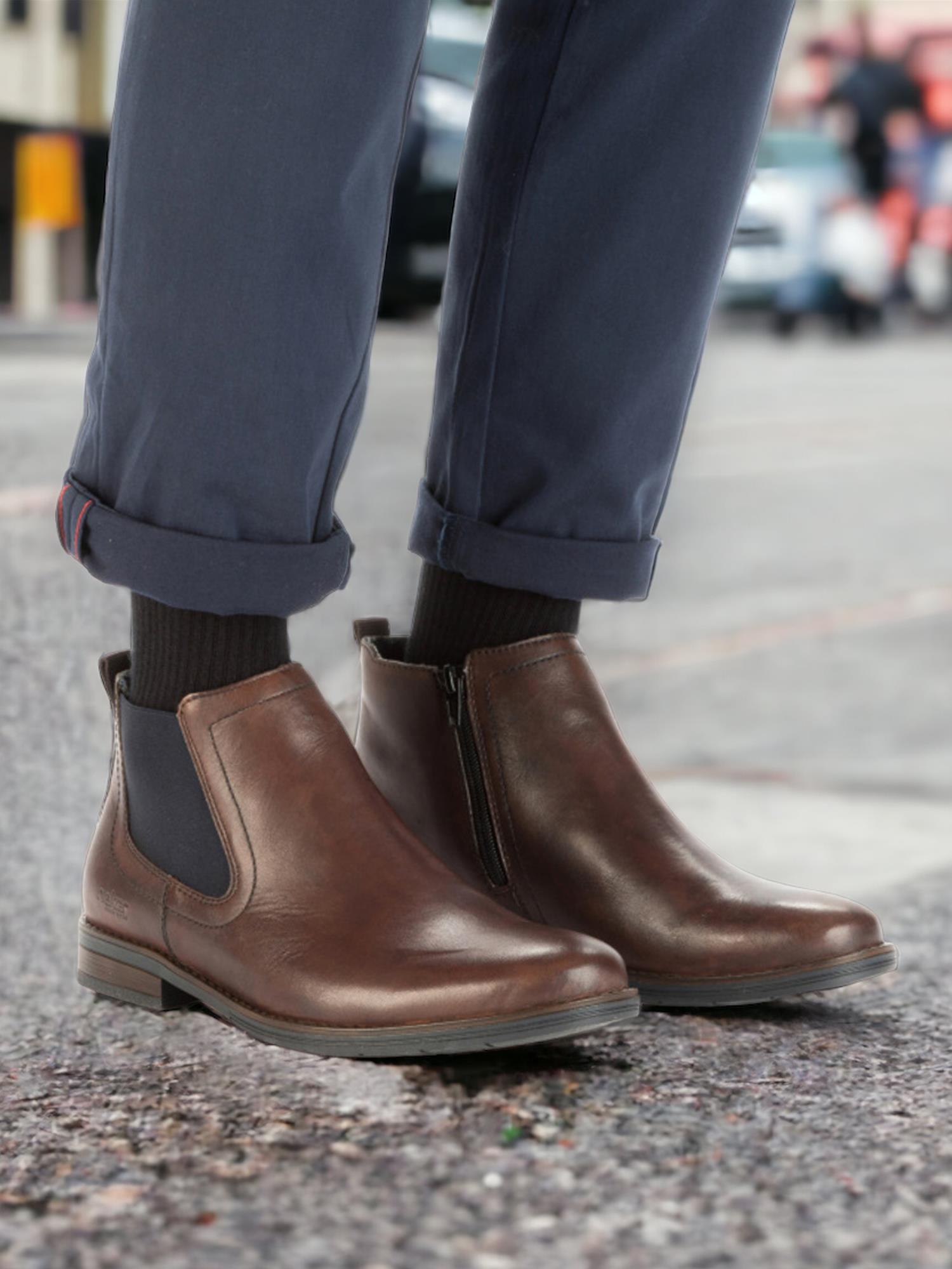 En person har på seg marineblå bukser med mansjett, svarte sokker og brune 04361 Rieker Testadimoro Short Boot Leather fra Rieker Comfort & Style med Antistress-teknologi, som sikrer komfortabel bruk hele dagen i en bygate.