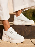 En person i hvite bukser med vide ben har på seg Rieker Comfort & Style 03280 Essential Comfort Sneakers med Antistress-teknologi, tykke teksturerte såler, hvite snørebånd og subtilt sidebranding mens hun står på en steintrapp utendørs.