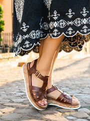 Nærbilde av en person iført Rieker Comfort & Style 03447 Toscana Leather Sandal med brune reimer og spenner, stående på en brosteinsbelagt gate. Et mørkt skjørt med hvite broderier fullfører stil og velvære-looken.