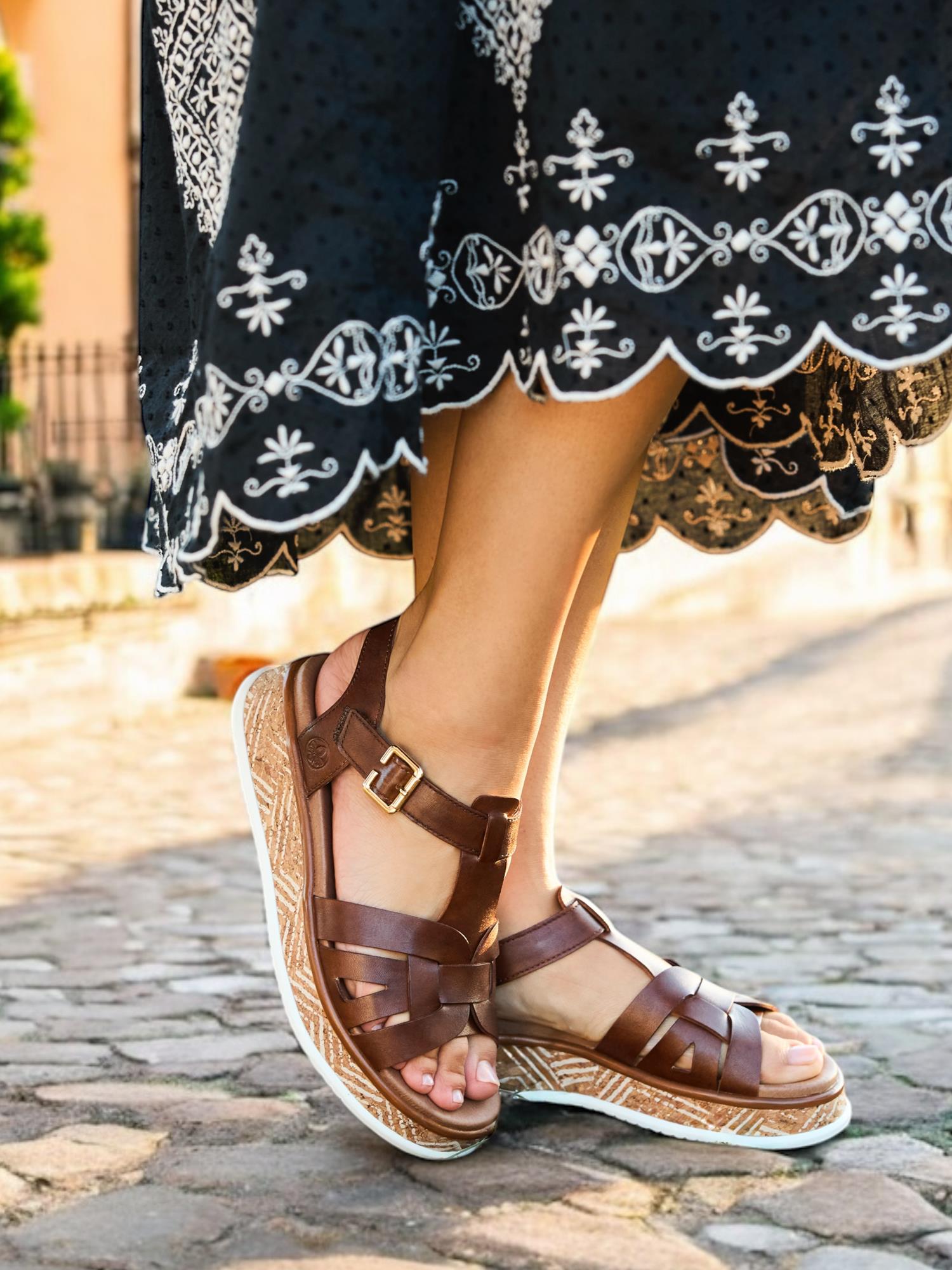 Nærbilde av en person iført Rieker Comfort & Style 03447 Toscana Leather Sandal med brune reimer og spenner, stående på en brosteinsbelagt gate. Et mørkt skjørt med hvite broderier fullfører stil og velvære-looken.